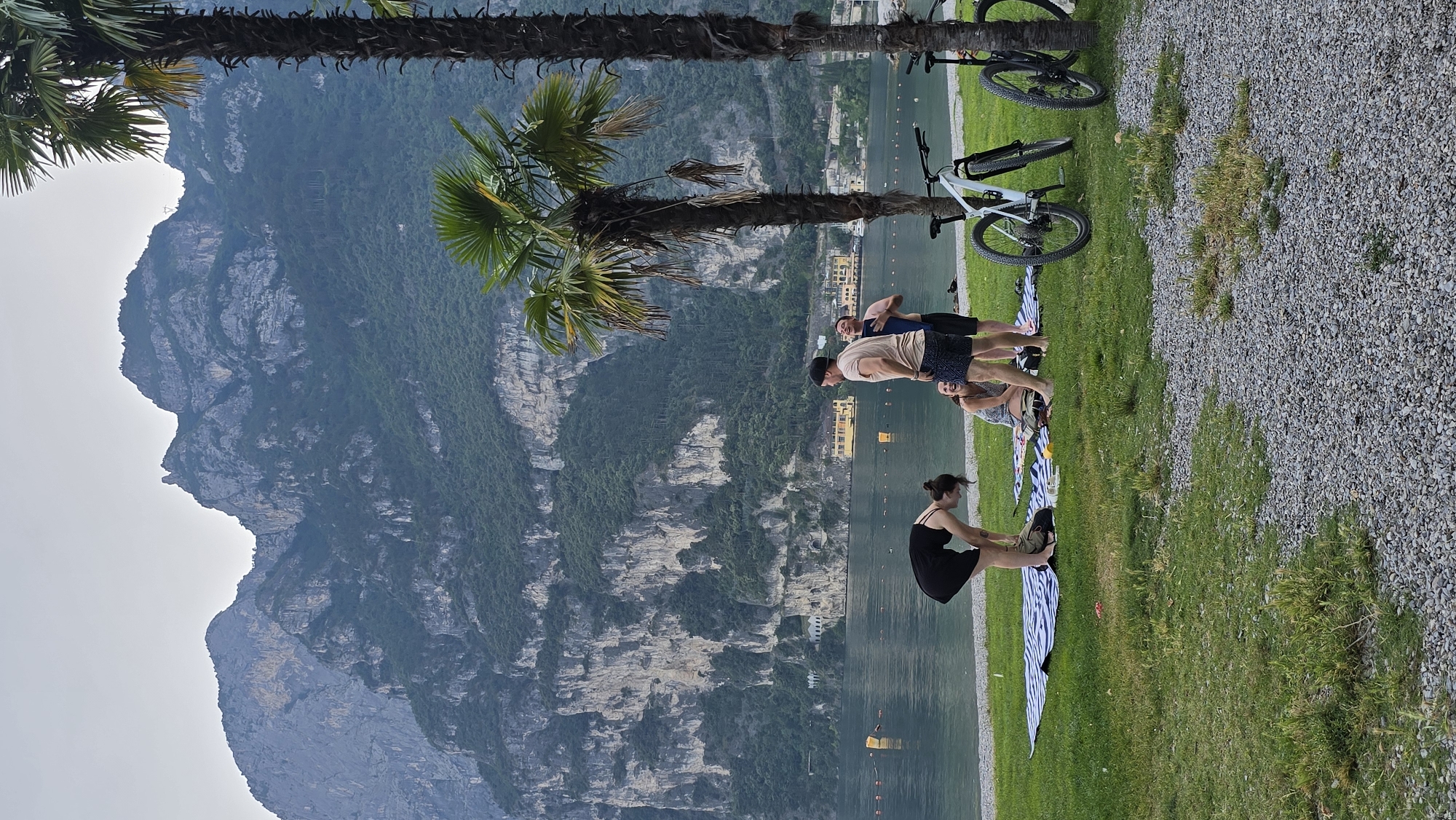 In Riva del Garda, Italy, a group of four people is gathered on a grassy area by a lake. A woman in a black dress is bending down, appearing to adjust her shoes or something on the ground. To her right, another woman is seated on a striped blanket, laughing. Two men stand to the right of the seated woman. One man, wearing a beige t-shirt and dark patterned shorts, is looking at the woman in the black dress. The other man, wearing a dark t-shirt and dark shorts, is looking away from the group and laughing. Two bicycles are parked on the right side of the frame, one white and one black. The background features a serene lake with buoys visible in the water, and beyond the lake, steep, tree-covered mountains rise under a hazy sky. The scene suggests a casual outdoor gathering or break, possibly after cycling.
