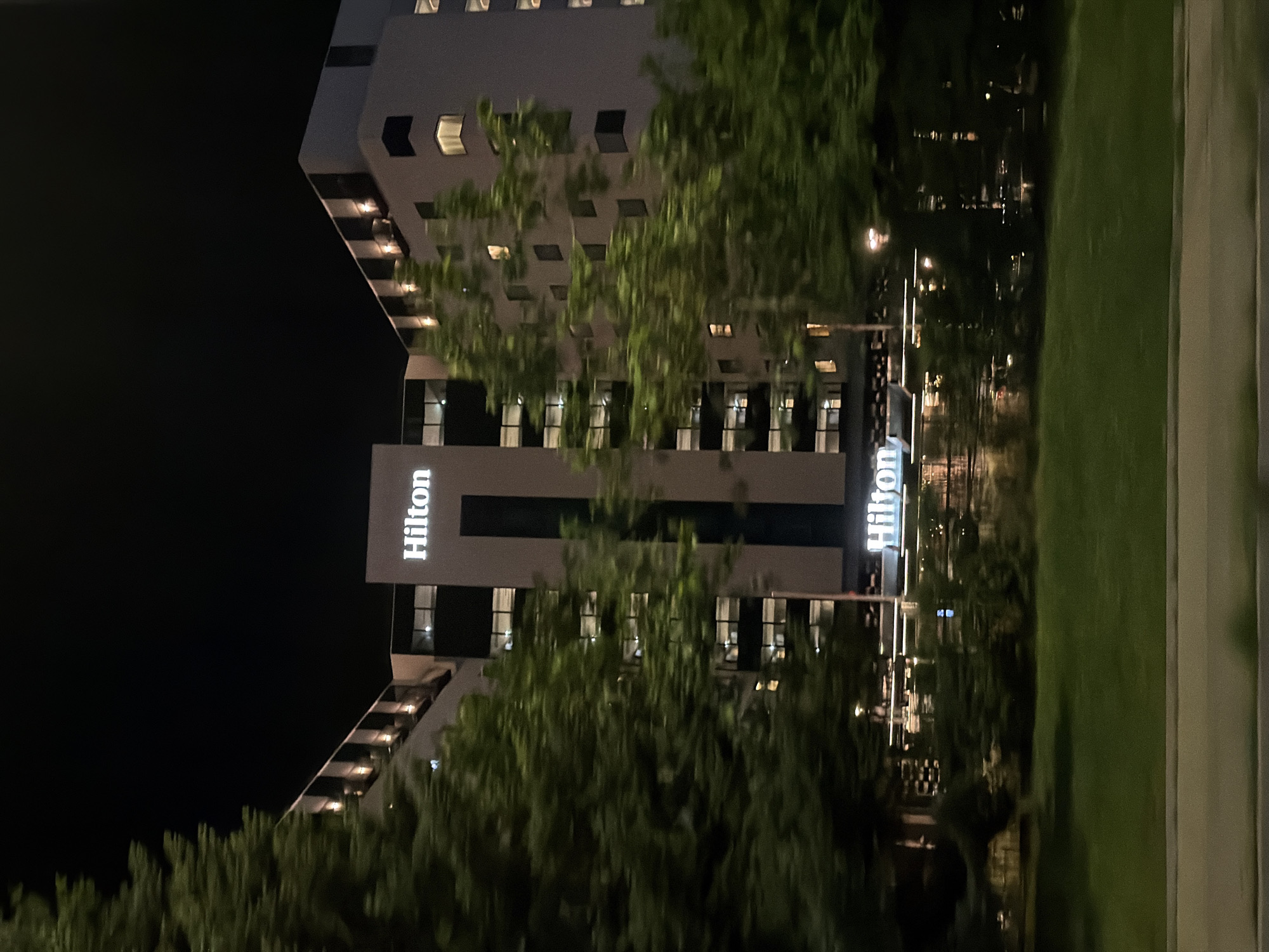 The image depicts the Hilton hotel in Sofia, Bulgaria at night. The hotel is a tall, modern building with a prominent illuminated "Hilton" sign. Trees with lush green foliage are in the foreground, partially obscuring the hotel's facade. The sky is dark, indicating nighttime. The image also shows a grassy area and a road in the lower portion, suggesting it was taken from a moving vehicle. The lighting from the hotel and surrounding areas creates a warm glow against the dark backdrop.