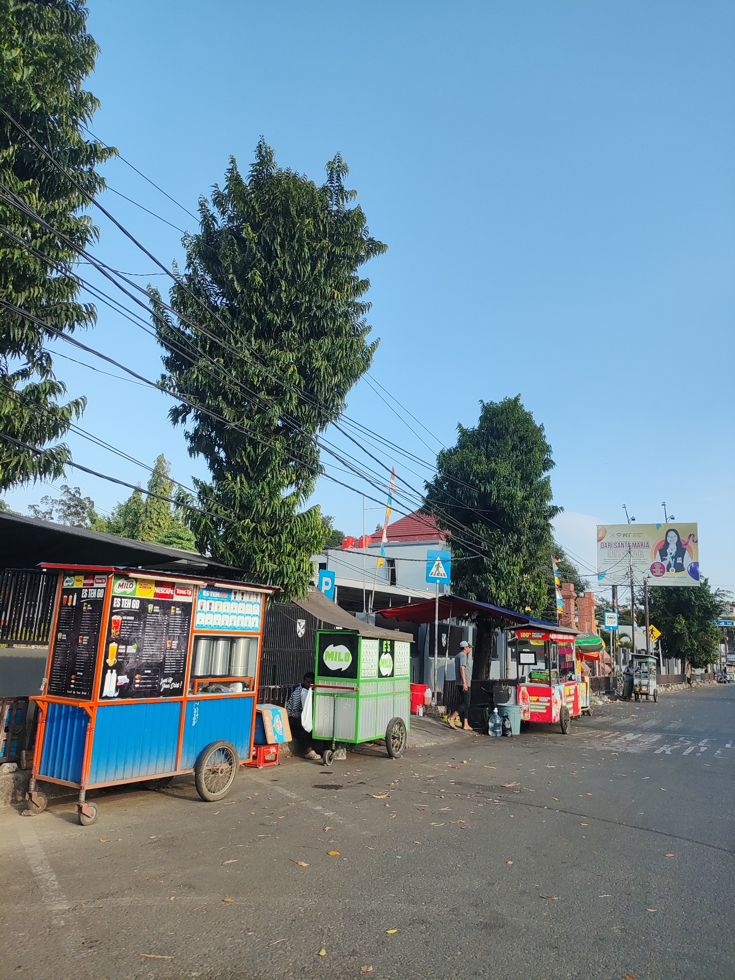 The image depicts a street scene in Cirebon, Indonesia, likely on a sidewalk adjacent to a road. The day is bright with a clear blue sky. The foreground is dominated by a blue and orange mobile food cart selling drinks, with various menu options displayed prominently. To the right of this cart is a green cart, also appearing to be a food or drink vendor, with the word "MILO" visible. Further down the street, other similar carts and small shops are visible, suggesting a commercial area.
Several tall trees with dense foliage line the street, their branches partially obscuring a network of overhead power lines. In the background, buildings with tiled roofs and a large billboard advertising "DARI SANTA MARIA UNIVERSITAS" are visible. A few people are present, including one individual standing near the green cart and another person further down the street near a different vendor. The road surface is asphalt, with fallen leaves scattered across it. The overall impression is a bustling, everyday street scene in an Indonesian city.