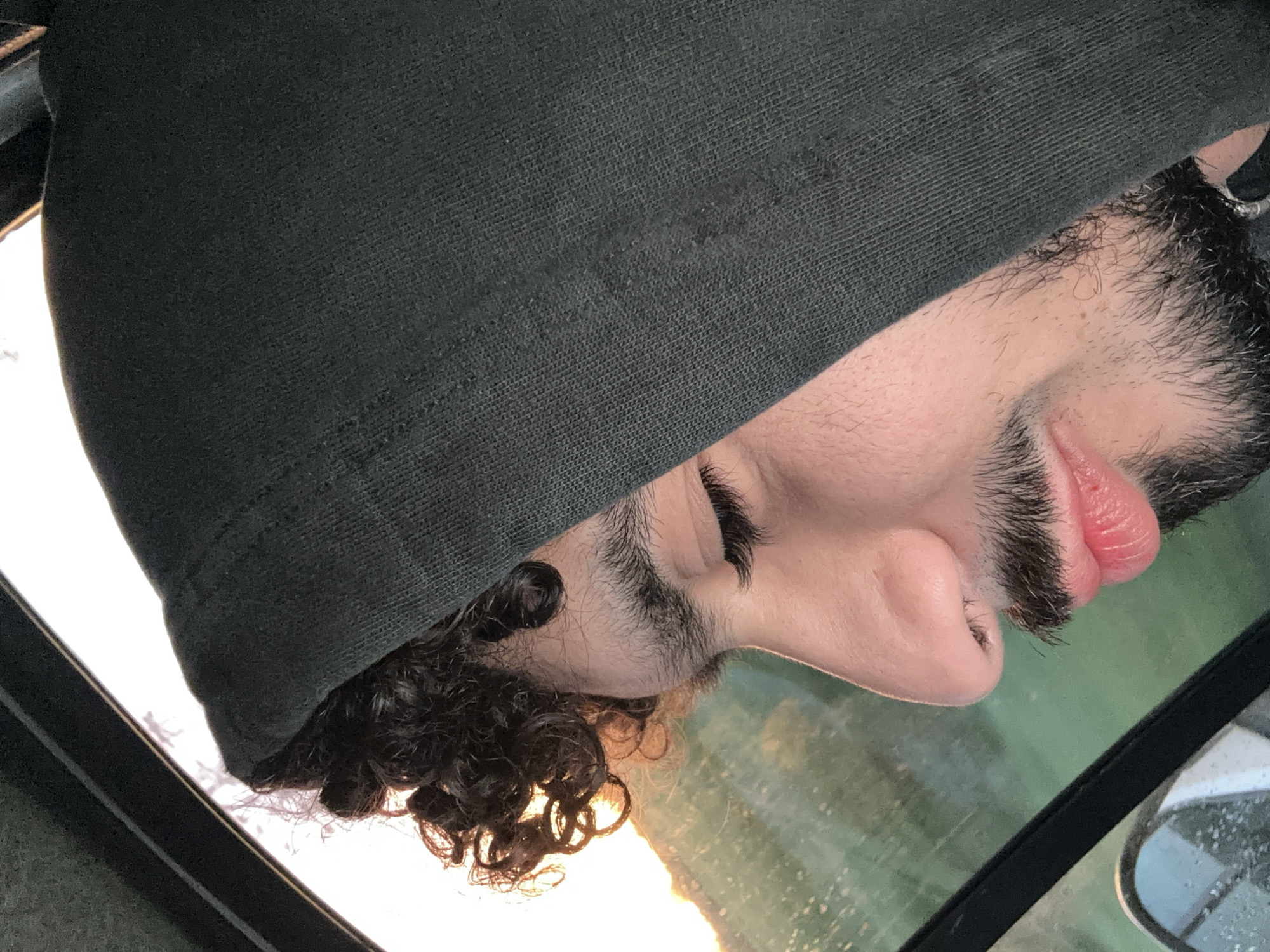A medium close-up, upward-angled shot shows a person's face partially covered by a dark hooded sweatshirt. The person has dark curly hair visible at the bottom left. The face, including the nose, closed eyes with dark eyelashes, and mouth with pink lips, are visible in the upper right area. The person appears to have facial hair. The scene is framed by a black, angled surface, suggesting the person is inside a vehicle, possibly a car, with the face reflected in the window. Light is visible on the lower side of the frame, suggesting a sunrise or sunset. The setting appears to be outdoors. There is no information from the picture to suggest it was taken in Villejust, France.