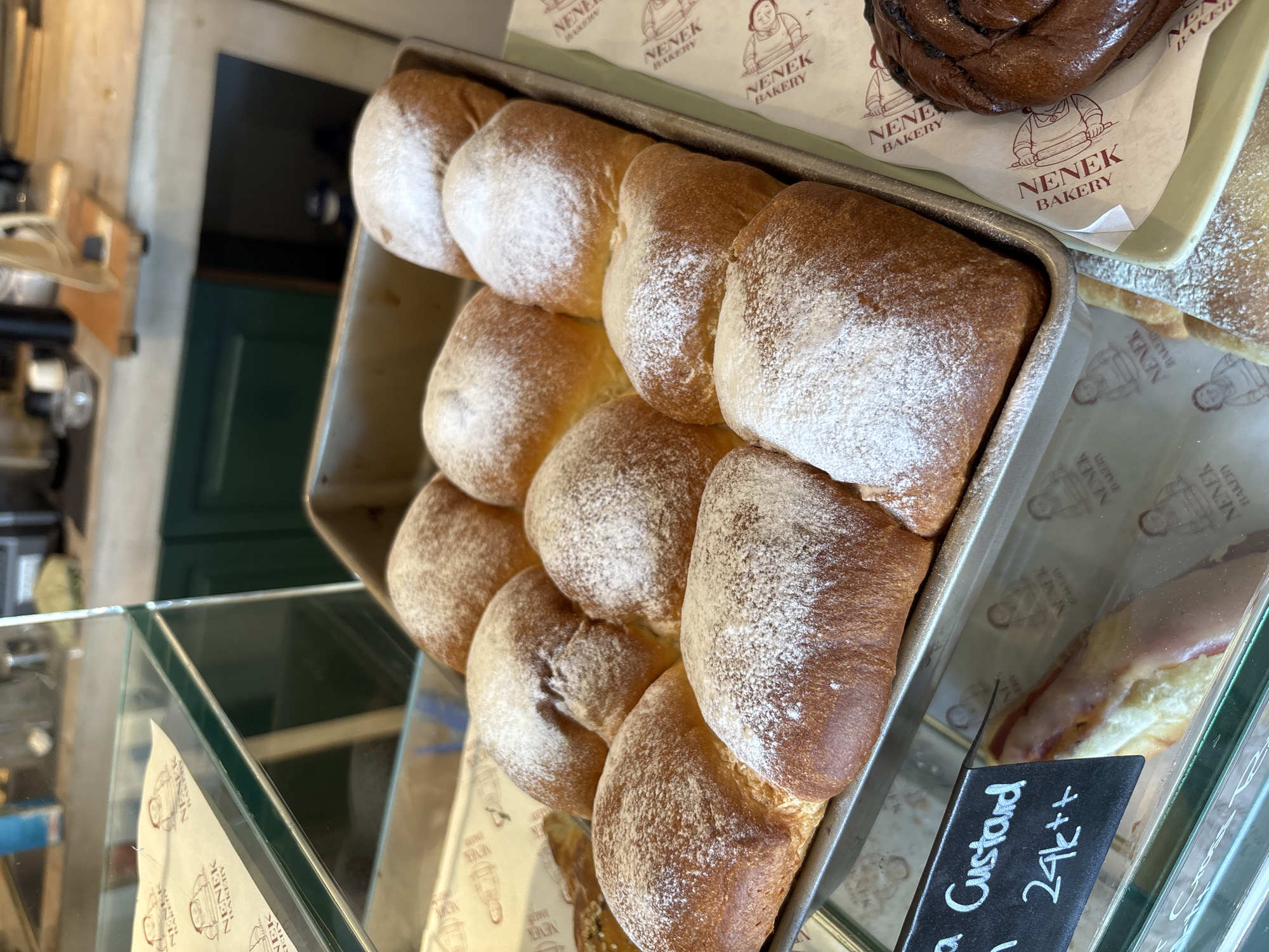 A close-up shot inside a bakery, possibly located in Ungasan, Indonesia, shows a metal baking pan filled with a row of golden-brown, square-shaped buns heavily dusted with powdered sugar. The buns are compactly arranged, filling most of the pan. Adjacent to this is a tray displaying a dark, twisted pastry, and a pastry with a light-colored glaze. The pastries sit atop paper printed with the name "NENEK BAKERY" and an illustration. A black sign with white writing is visible, with "Custard" and "24k++" legible, suggesting a menu item and price. The pastries are behind a glass display case. The background shows a glimpse of the bakery's interior, including a green door and a blurred view of equipment.
