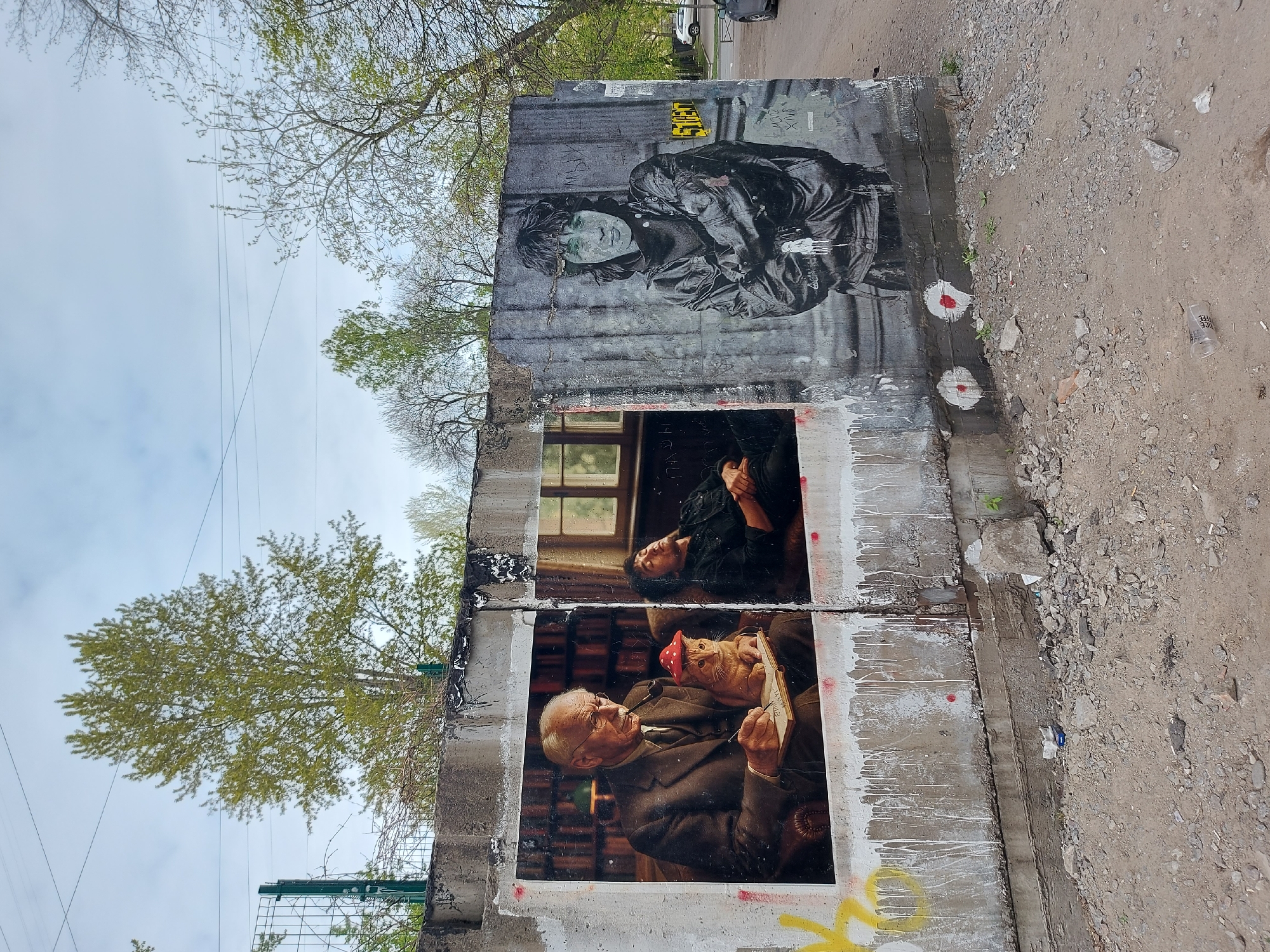 The image shows a dilapidated concrete wall with various artworks and graffiti in Saint Petersburg, Russia. The wall is partially destroyed, with visible signs of decay and weathering.
The upper section of the wall features a monochrome portrait of a person wearing a jacket, possibly a depiction of Viktor Tsoi. The middle and lower sections contain large, framed photographs or prints. One features a portrait of Carl Jung holding a pipe, with a person wearing a red mushroom hat. The other photograph depicts a man with dark hair lying with his head supported by his hand.
The wall is splattered with white paint drips and has graffiti in yellow and other colors. The surrounding area consists of dirt, gravel, and sparse vegetation. Trees with green foliage are visible in the background. Overhead power lines are also present.
