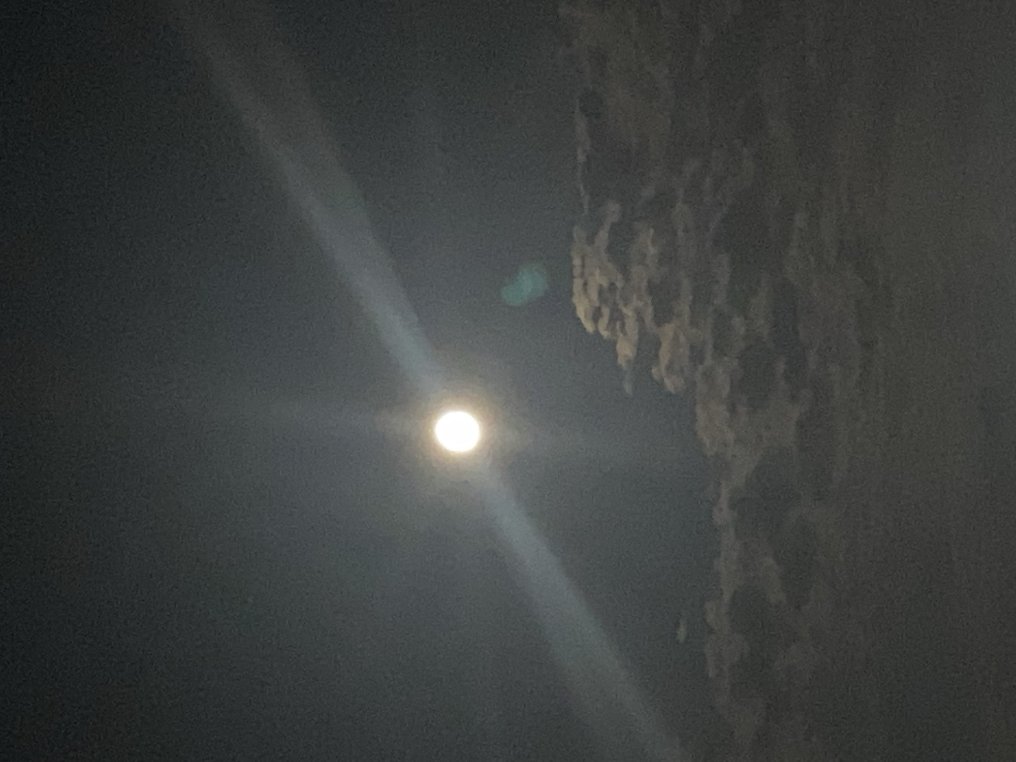 The image depicts a nighttime sky with a bright full moon. A distinct beam of light emanates from the moon and extends diagonally across the frame. Scattered clouds are visible in the lower portion of the image, illuminated by the moonlight. A subtle green lens flare is present to the right of the moon. The setting appears to be an outdoor environment within the city of Iași, Romania, likely a place with an unobstructed view of the night sky. There are no people or specific urban structures clearly identifiable in the image.