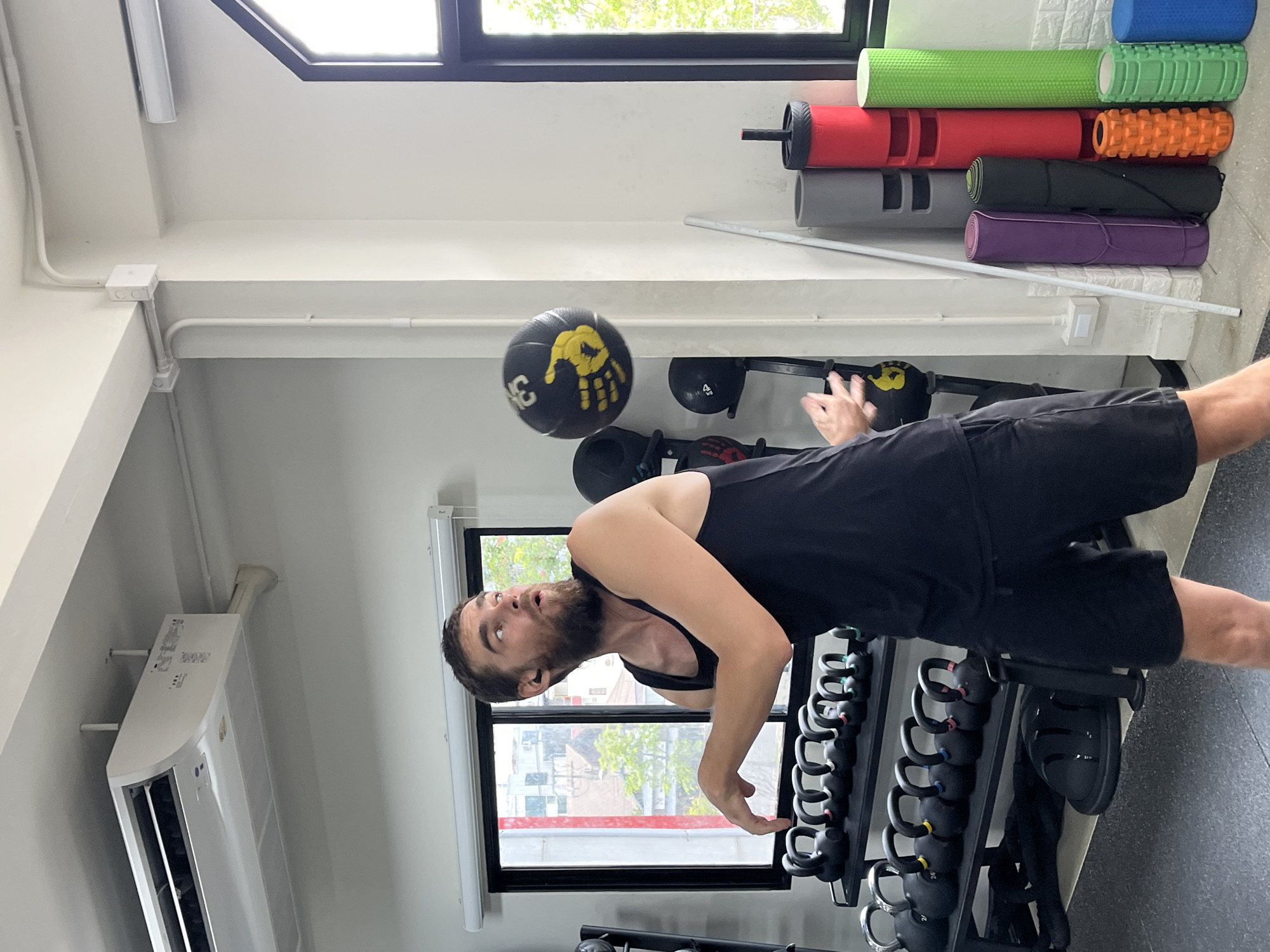 A Caucasian male with a beard is captured in mid-action at a gym in Chiang Mai City Municipality, Thailand. He is wearing a black tank top and black shorts, and his right arm is extended as if he has just thrown or is about to catch a black medicine ball with yellow markings that is suspended in the air to his right. His gaze is directed upwards, following the trajectory of the ball. The background reveals a typical gym environment: a rack of black kettlebells to the left, and various exercise equipment including foam rollers, yoga mats, and a medicine ball holder on the right. An air conditioning unit is visible on the ceiling. The scene is lit by natural light filtering through a large window in the background, which offers a view of buildings and greenery, suggesting an urban setting. The overall impression is of a dynamic fitness activity taking place in a well-equipped gym.