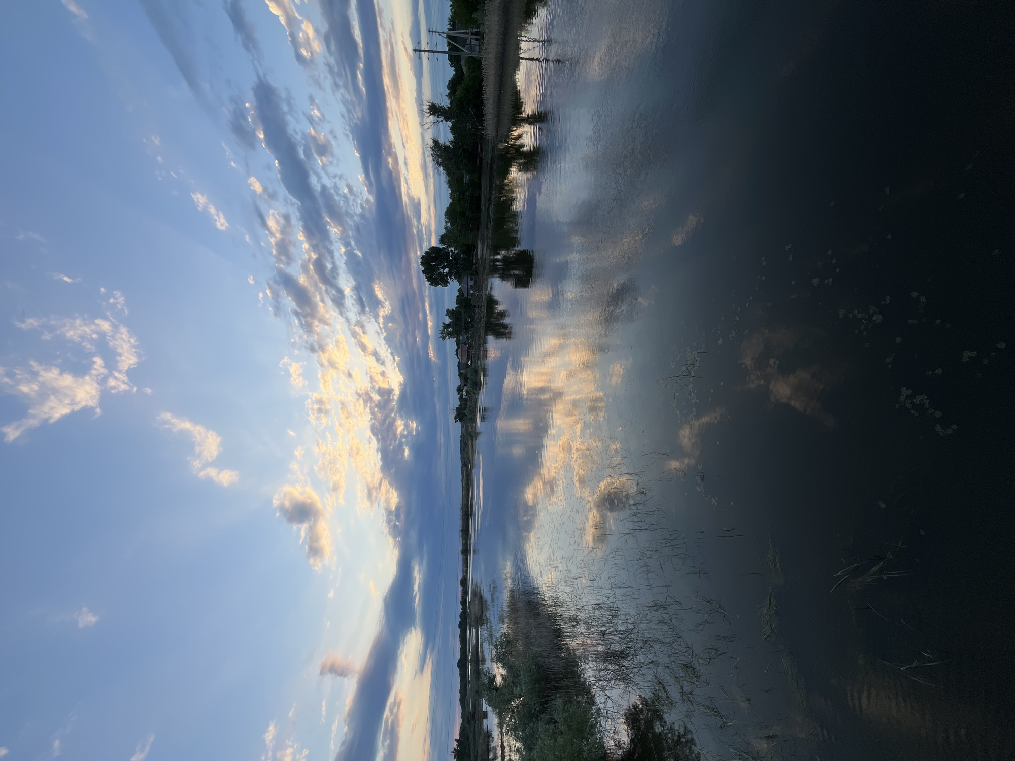 This is a vertical, wide-angle shot of a body of water at dusk in Krasnyye Barrikady, Russia. The sky is a soft blue with scattered white and gray clouds, some illuminated by the setting sun, casting long rays of light. The water's surface is calm and reflective, mirroring the sky and the trees and structures along the far bank. The left side of the image shows the sky in detail, with distinct cloud formations and sunbeams. The right side is dominated by the dark, reflective water, with some subtle patterns of aquatic vegetation visible. Along the distant horizon, a line of trees and a few buildings are visible, along with a utility pole. The overall mood is tranquil and serene, characteristic of a peaceful evening in a rural setting. There is no visible text in the image.