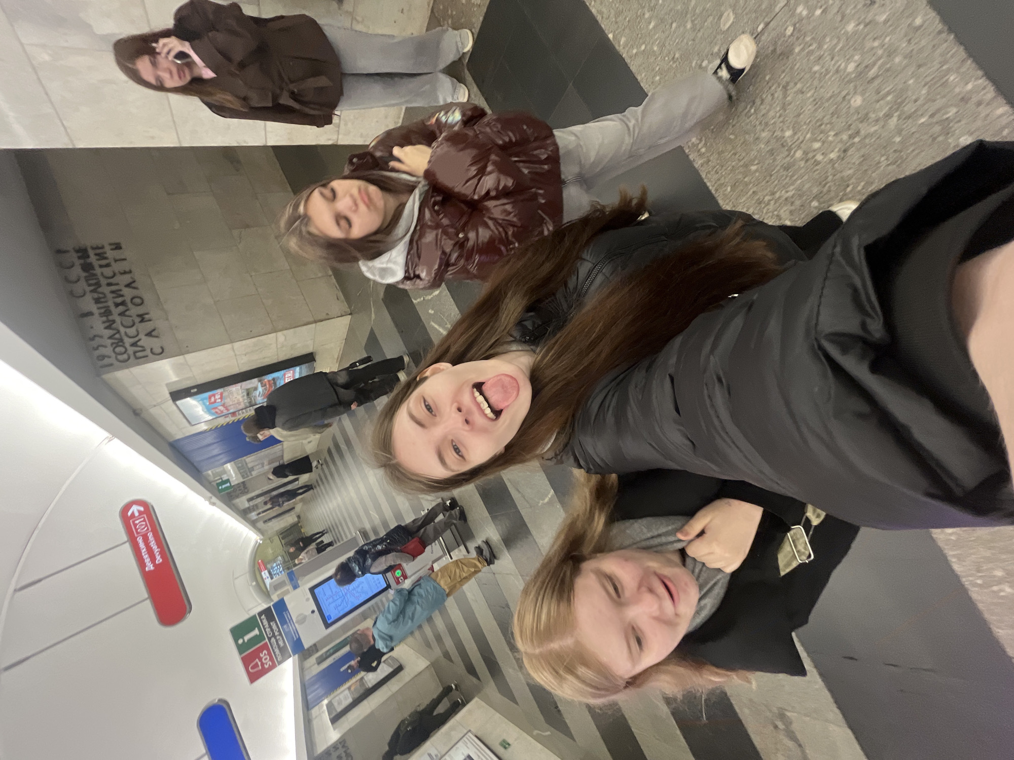The image depicts a group of four young women in what appears to be a subway station in Saint Petersburg, Russia. One woman is speaking on her cell phone, another has her eyes closed, one is sticking her tongue out, and the other is smiling. The subway station has signs in both Russian and English. The signs indicate this location is the Devyatkino subway station. The subway platform is crowded with people.