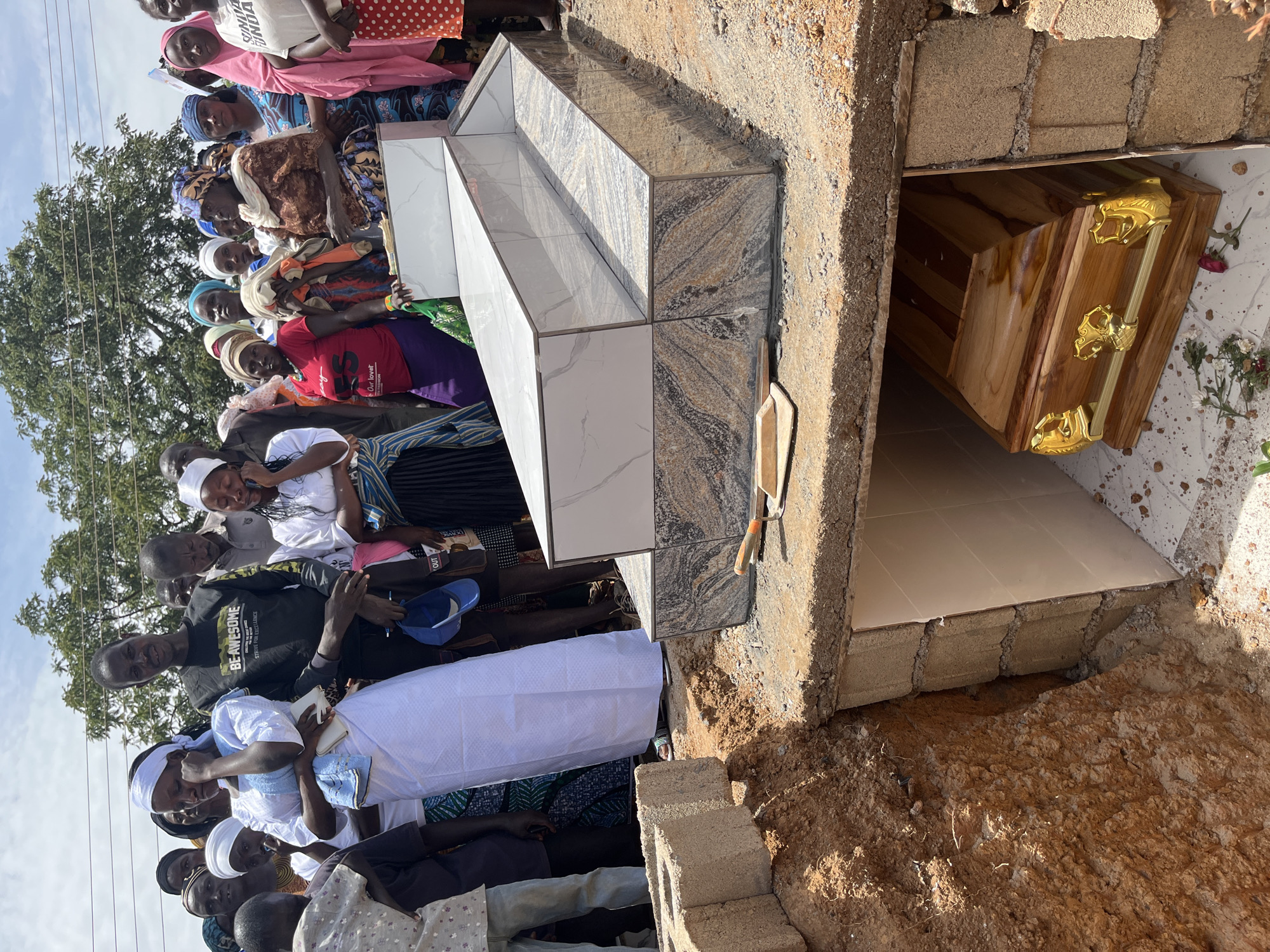 A funeral is taking place in Dogon Dinia, Nigeria. A crowd of people surrounds a grave. A wooden coffin with gold-colored handles sits in the grave. The grave is lined with concrete blocks and partially covered with tile. Some of the people appear to be grieving.