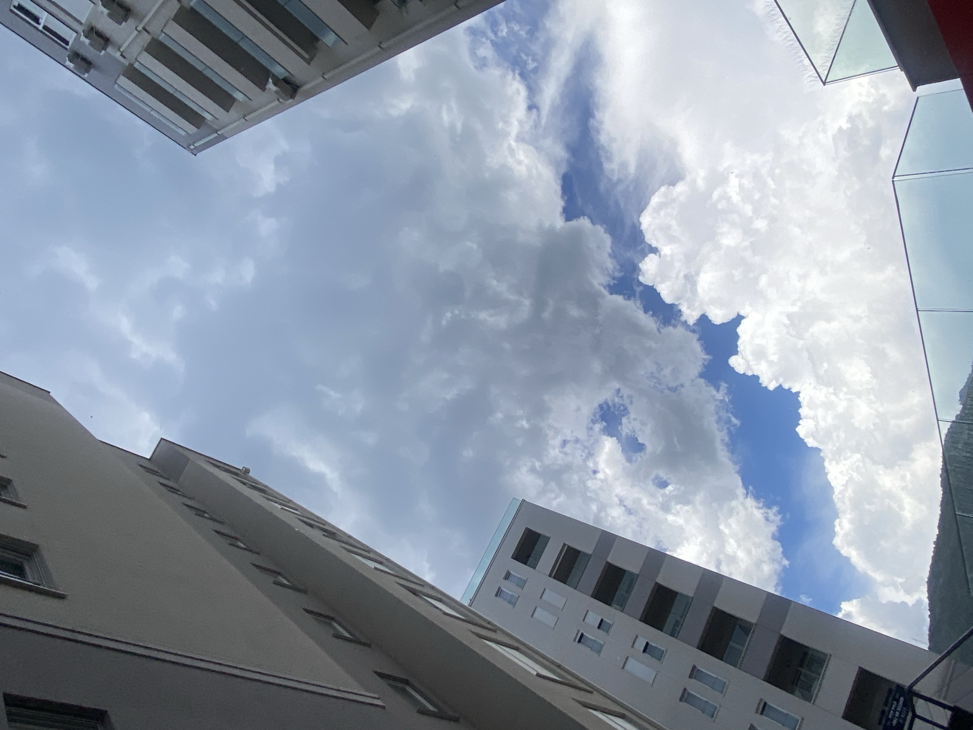 This media file is a low-angle shot looking up at buildings and the sky. The scene is an urban setting, likely a city, with modern architecture. Three distinct buildings are visible, their facades angled towards the viewer. The building in the foreground on the left is a light-colored, multi-story structure with numerous windows. Another building, on the right and further back, is also tall and appears to have a mix of dark-framed windows and lighter sections. A third building, at the top of the frame, is partially visible and shows a modern design with horizontal bands.
The sky is filled with dynamic clouds. Large, fluffy white cumulus clouds dominate the upper right and center of the frame, interspersed with patches of bright blue sky. The clouds suggest a partly cloudy day, possibly with the possibility of rain or recent rain, given the varied cloud formations. The lighting indicates it is daytime, likely late morning or afternoon, as the sun is illuminating the clouds brightly.
There are no people visible in the media file. The overall mood conveyed is one of looking up and experiencing the scale of the buildings against the vastness of the sky. The location context provided is Budva, Montenegro, which suggests a coastal city or a city with significant modern development. No visible text is present in the image.