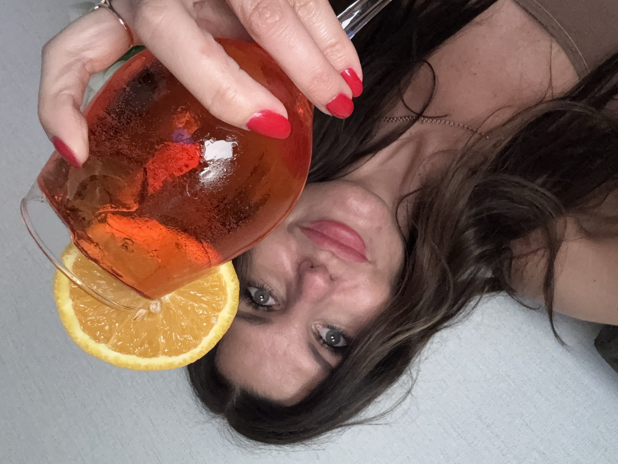 A woman with long, dark, wavy hair and grey eyes holds a clear glass filled with an orange-red beverage topped with ice and a slice of orange. She is wearing a brown, strapless top and a delicate chain necklace. Her fingernails are painted bright red. The woman's face is partially visible as she takes a selfie, holding the drink up towards the camera. The background appears to be a lightly textured, pale wall. The image was taken indoors, possibly in a residential setting. There is no information to suggest this image was taken in Chaikovsky, Russia.