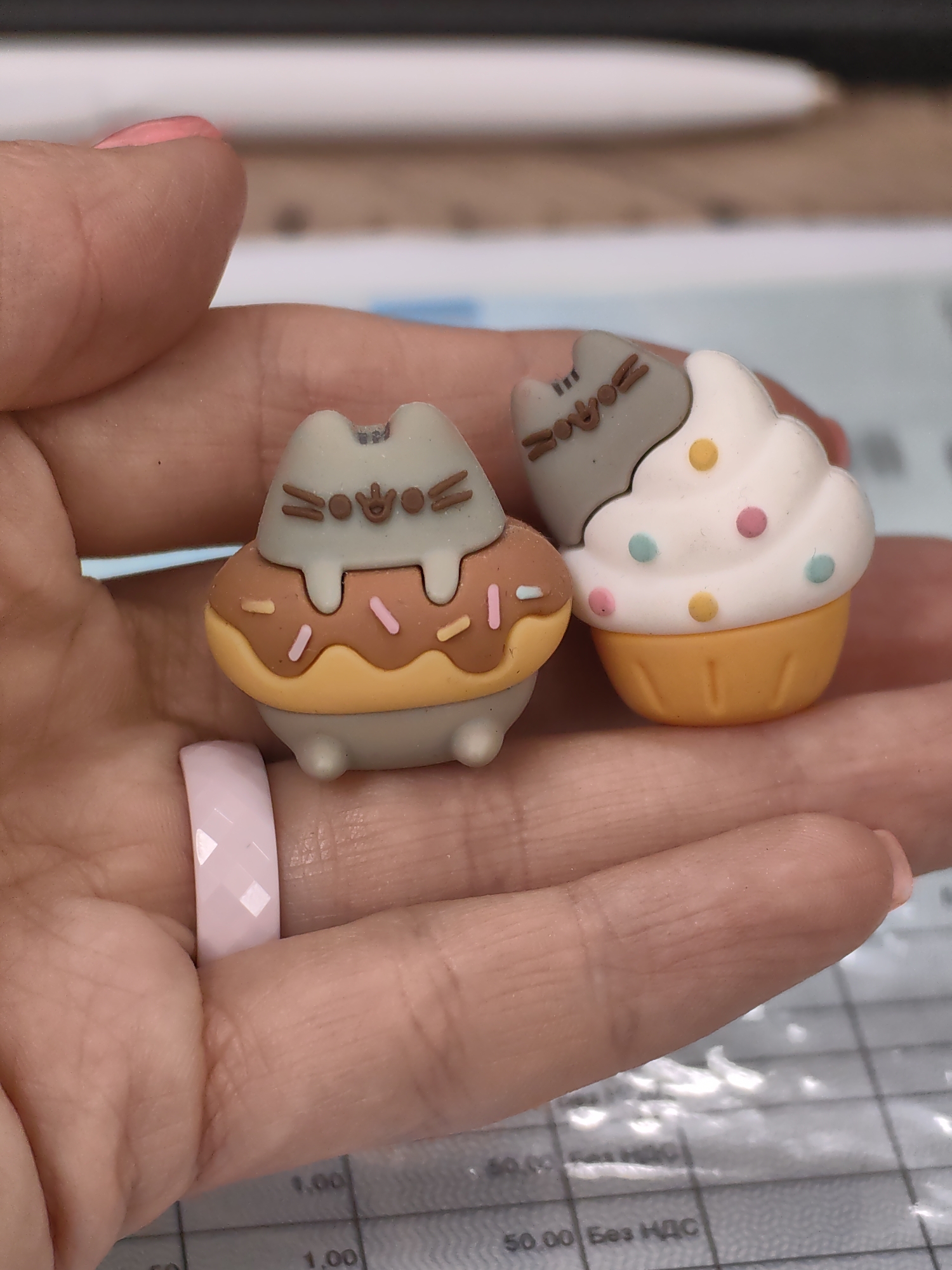 A close-up, eye-level shot shows a person's left hand holding two small, novelty erasers shaped like Pusheen the cat. The eraser on the left is gray and resembles a donut with brown icing and pink and orange sprinkles. The eraser on the right is white and shaped like a cupcake with colorful dots. A light pink ring with a faceted design is worn on the ring finger of the hand. The background appears to be a desk with a grid pattern, possibly a cutting mat, and some papers. The scene is not indicative of any specific location in Minsk, Belarus, or any other city.