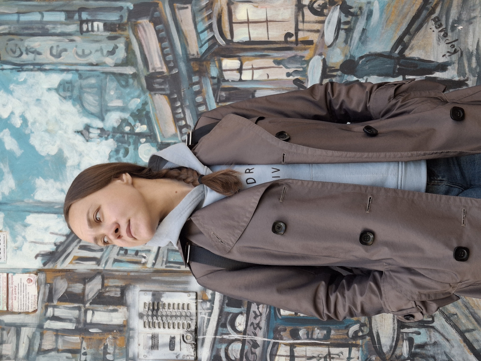 A woman with a braided ponytail and wearing a grey hooded sweatshirt and brown trench coat poses against a painted mural. The mural is an urban scene, depicting buildings, windows, a cloudy sky, and what appears to be a payphone or communication device. The name "Chaikovsky" is legible on a painted sign within the mural. The image orientation is sideways.