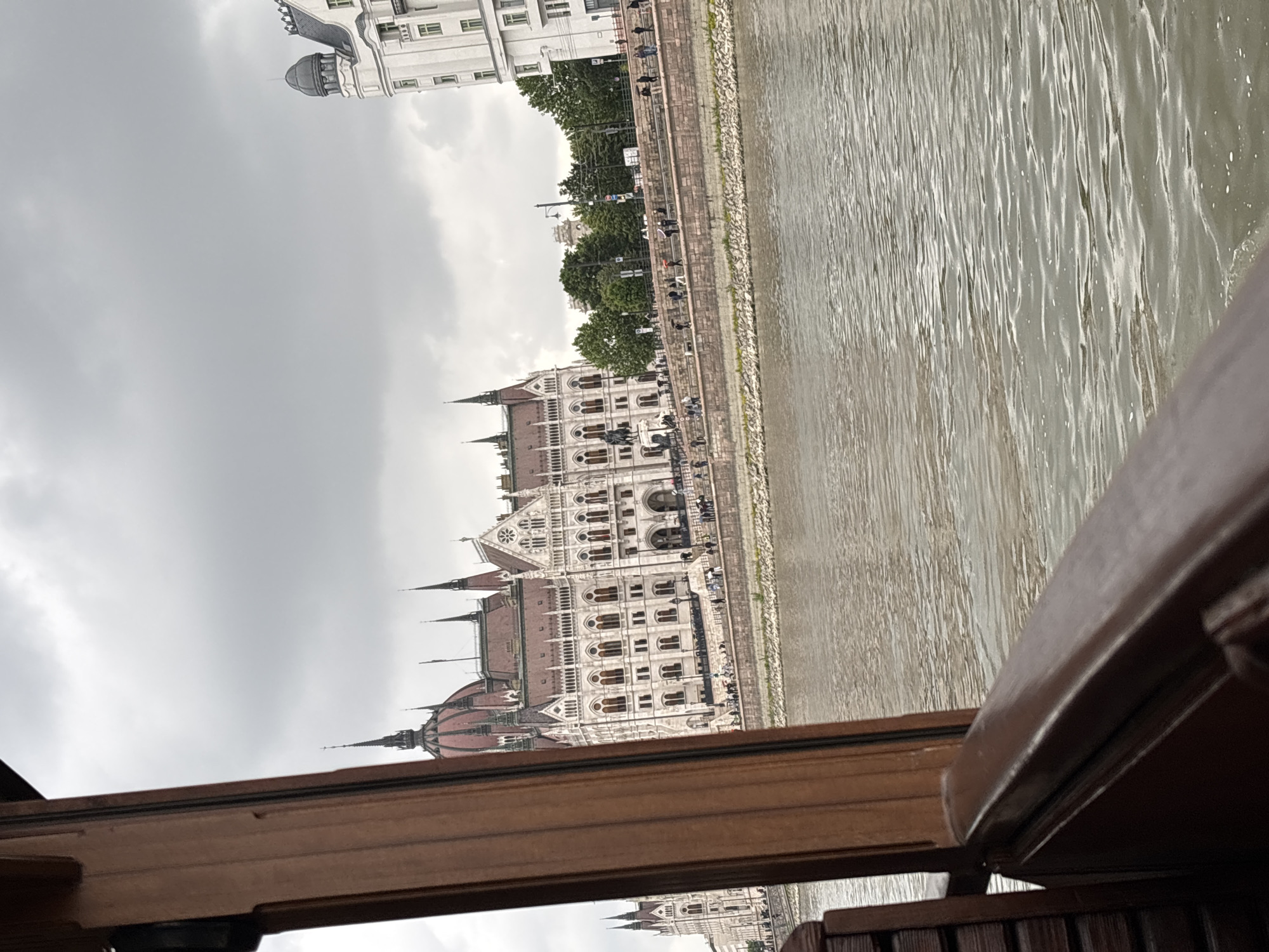 The image shows the Hungarian Parliament Building in Budapest, Hungary, viewed from a boat on the Danube River. The river fills the right side of the frame, displaying the water’s surface, while the parliament building occupies the center-left, showing its architectural details. A section of what appears to be the boat’s window frame and possibly the interior edge of the boat is visible along the bottom. The sky overhead is overcast. People can be seen on the embankment next to the building.