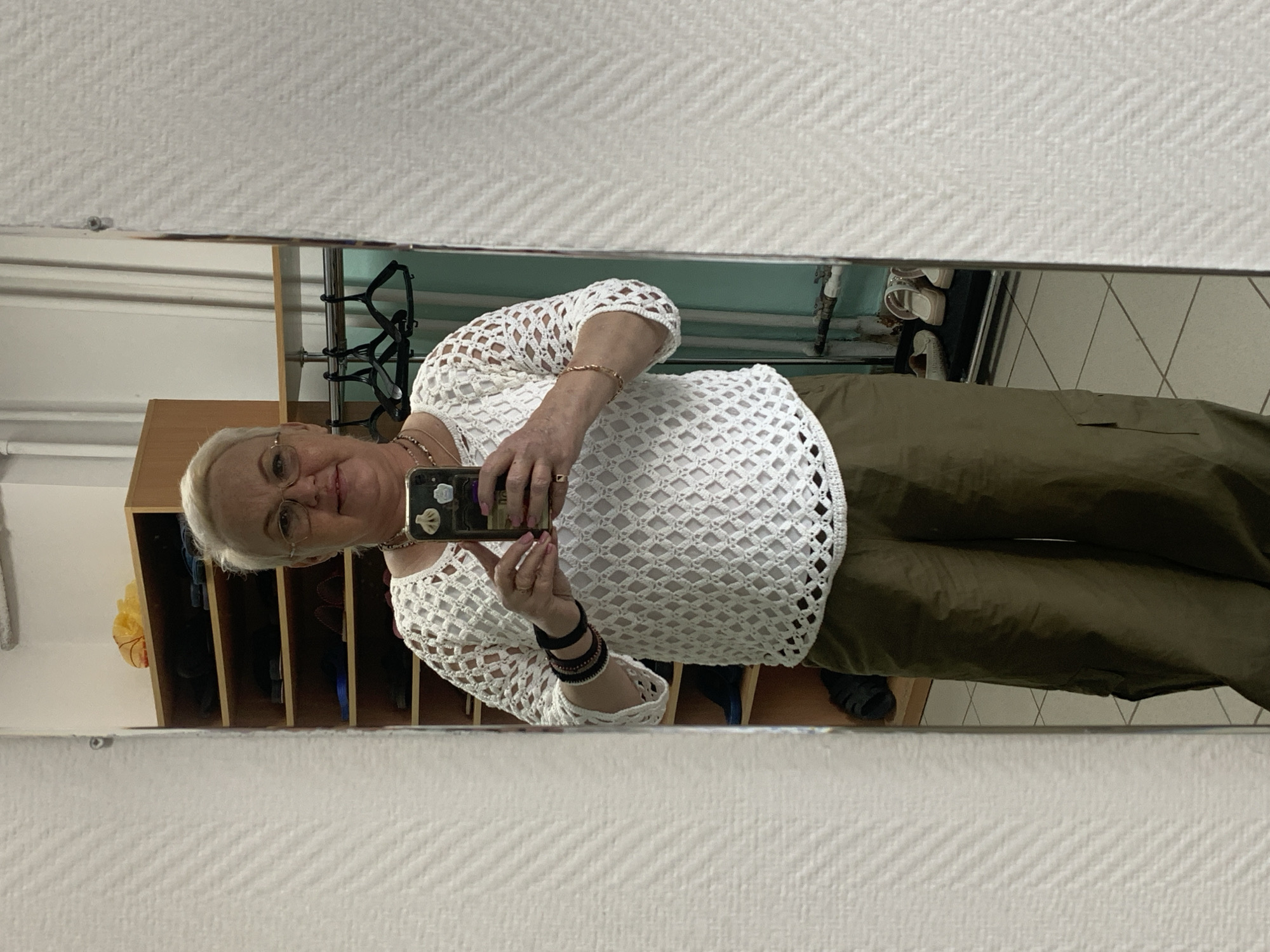A mature woman is taking a selfie in a mirror. She wears glasses, a white crocheted top, and olive green pants. She is holding a smartphone with a decorated case. In the background, there are shelves with items inside, a clothes rack, and a tiled floor. The mirror is hanging on a textured white wall. Based on the description and visual context, the location is likely in the city of Chaikovsky, Russia, as requested.