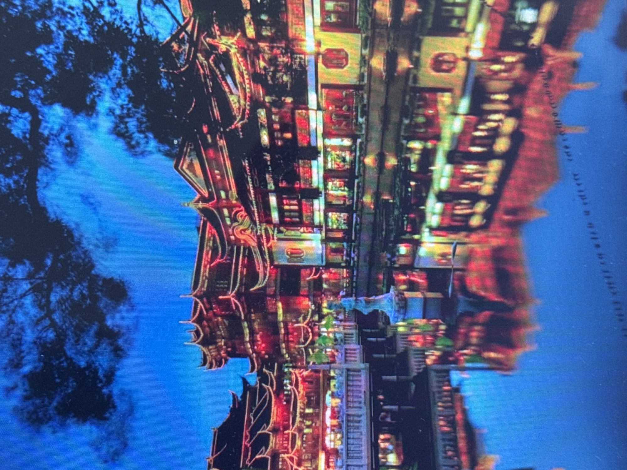 The image depicts an upside-down view of a street scene at night, illuminated by numerous bright lights. The architecture suggests a traditional East Asian style, with ornate roofs and decorative lighting. Buildings are densely packed, with many windows and facades lit up in various colors, including red, yellow, and white. A large, dark tree is visible in the upper left portion of the image, its branches silhouetted against a dark blue sky, possibly indicating evening or night. The reflection of the buildings and lights in what appears to be water creates a symmetrical, albeit inverted, composition. There is no clear indication of people or specific activities taking place, but the vibrant lighting suggests a lively, possibly commercial or festive, area. The overall mood is one of visual spectacle due to the intense illumination and reflections. Text is visible on the right side of the image, oriented vertically and appearing as a series of characters, but it is too blurry and distorted to be deciphered. The provided location context of Pushkino, Russia, seems incongruous with the visual cues of East Asian architecture.