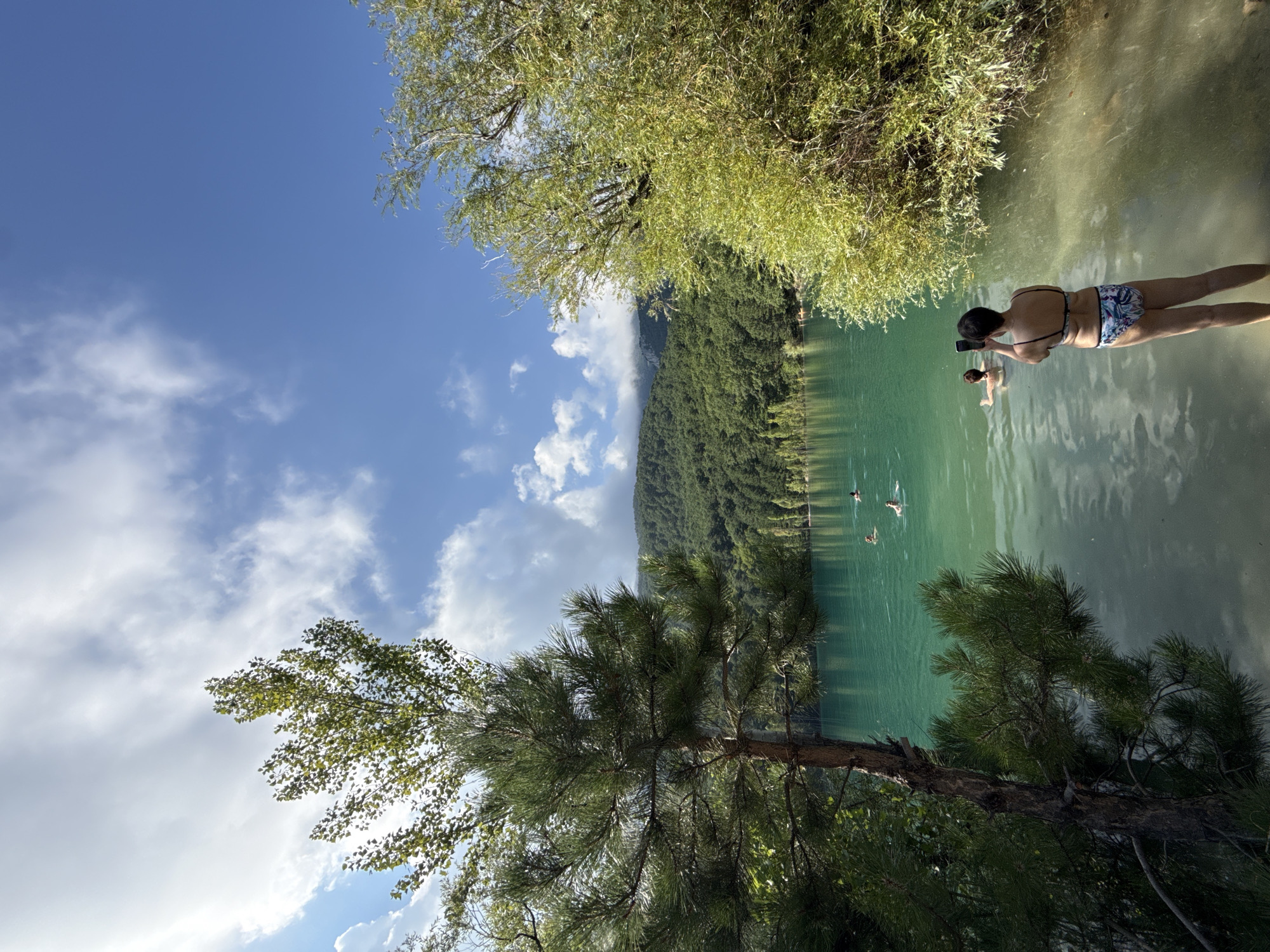 The image depicts a tranquil lake scene with a person in the foreground standing in shallow water and taking a photo with their phone. The person is wearing a bikini and facing away from the camera towards the lake. In the middle ground, several individuals are swimming in the clear, turquoise water. The background features a lush, tree-covered hillside under a blue sky with scattered white clouds. The environment is natural and serene, with no urban elements visible. The location is specified as Unknown, Ukraine.