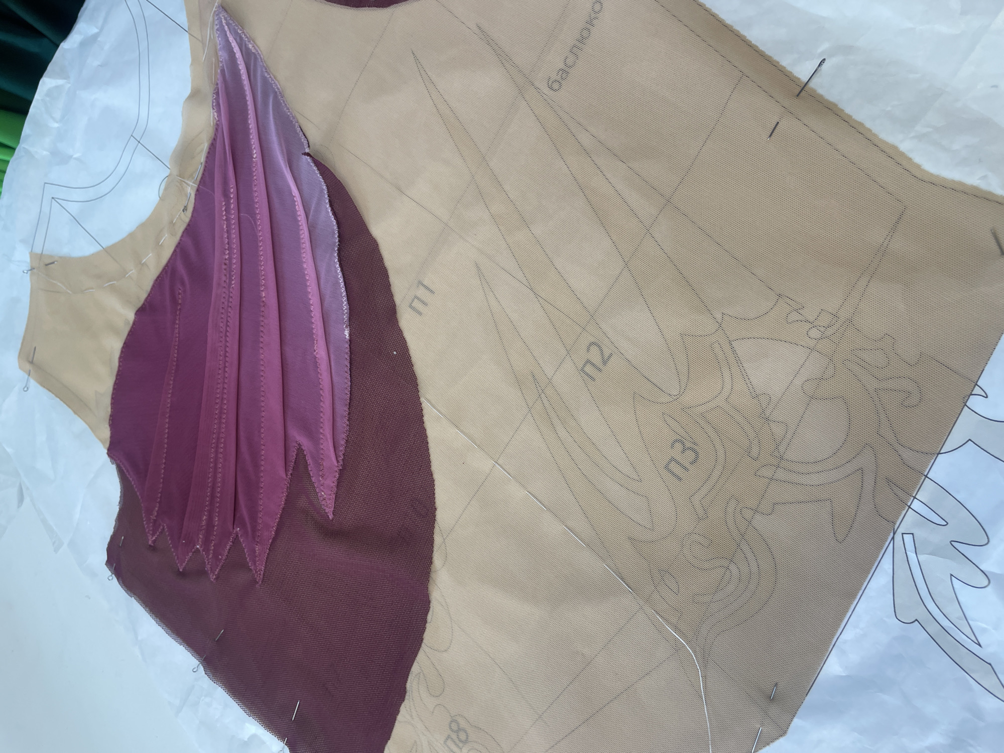The image shows a close-up view of a sewing project in progress. A tan-colored fabric is laid out on a white surface, with a sewing pattern underneath. Over the tan fabric, there are pieces of dark red and pink sheer fabric, layered and pinned in place. The pink fabric is pleated, creating a fan-like design. Sewing pins are visible, holding the fabric pieces together. The tan fabric has black lines of a sewing pattern printed on it, with some sections labeled with Russian letters and numbers, such as "п1", "п2", "п3" and "баслюкою". The overall scene suggests a creative endeavor, likely the making of a garment or costume, with an emphasis on intricate detail. There is no discernible information in the image to suggest a specific location in Saint Petersburg, Russia.