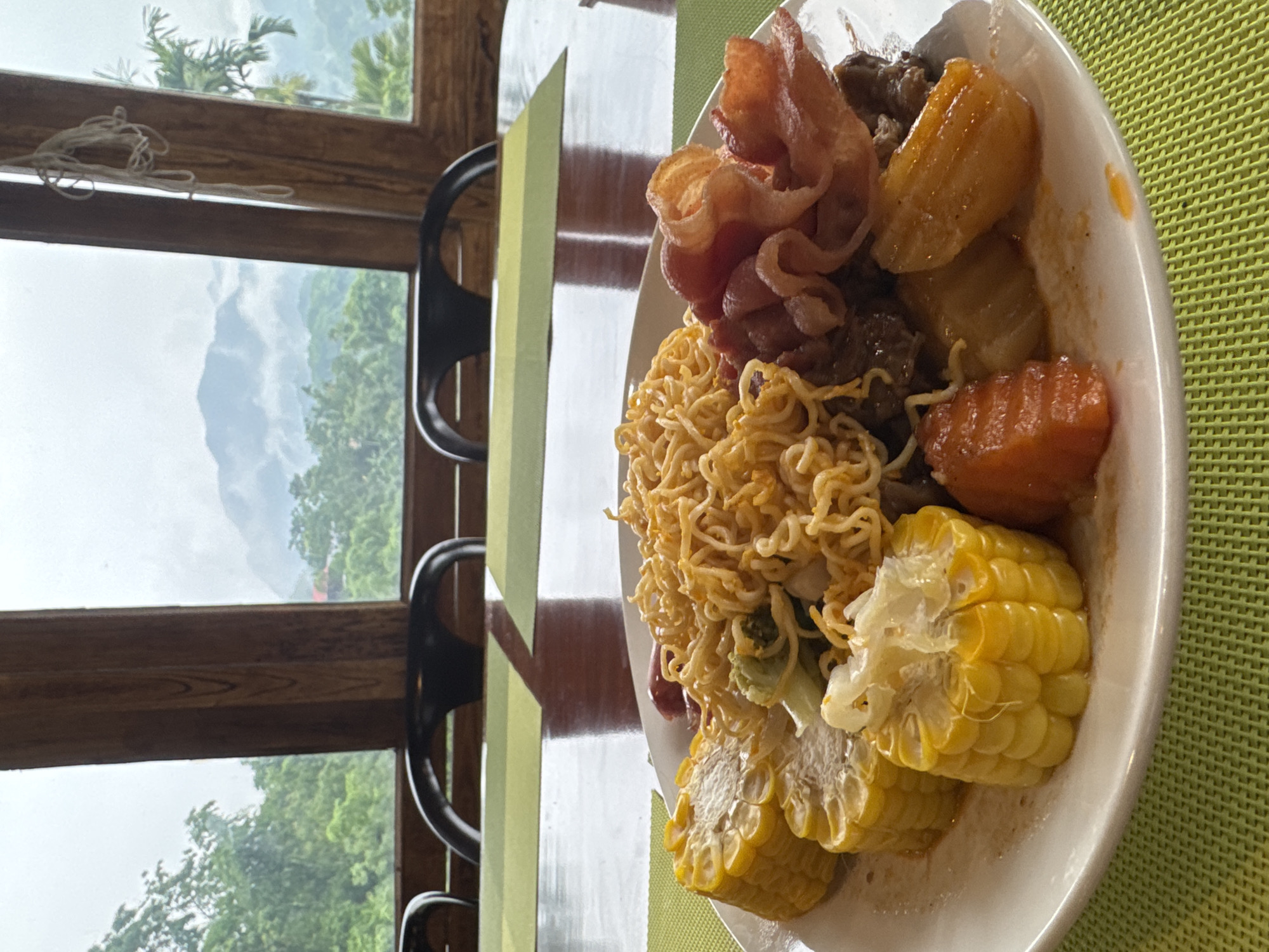 In Pù Luông Commune, Vietnam, a white plate filled with food rests on a green placemat. The plate contains corn on the cob, instant noodles, bacon, and what appear to be pieces of beef and carrots. In the background, through a large window with wooden framing, a misty mountain landscape with lush green trees is visible. The room appears to be a dining area with chairs and tables, suggesting a restaurant or lodging with a view. The atmosphere seems calm and possibly overcast due to the mist.