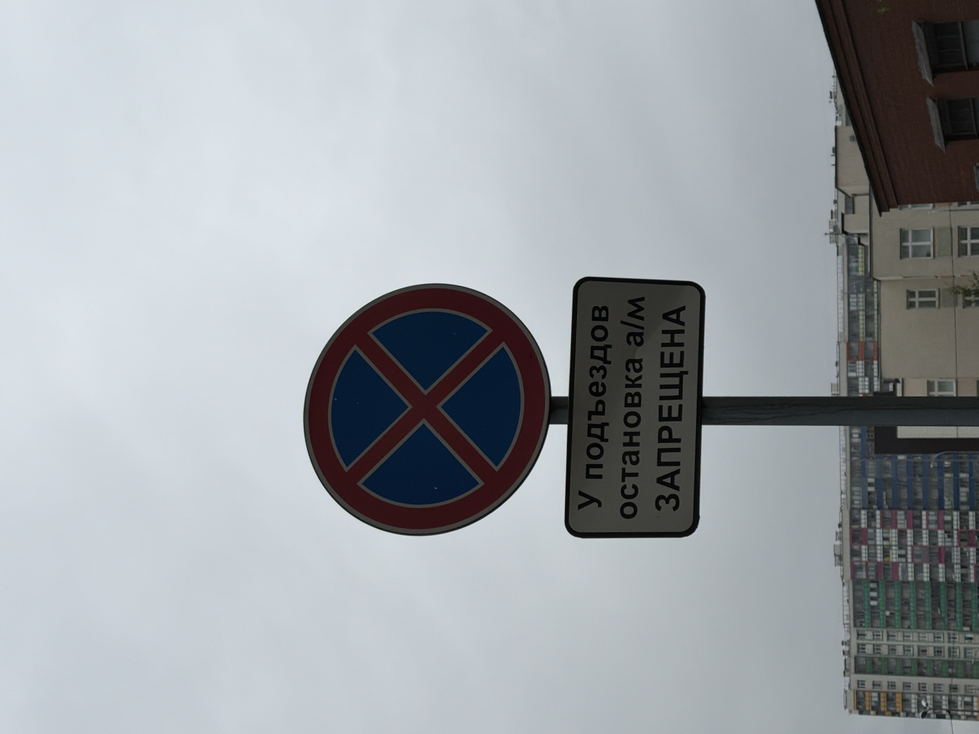 A “No Stopping” sign, featuring a red circle around a blue X, is positioned to the left of a white sign with black Cyrillic text, which reads “У подъездов остановка а/м ЗАПРЕЩЕНА” meaning “No stopping of cars at the entrances.” The sky is overcast and grey. Buildings are located in the background, including a predominantly beige structure with a brown roof at the upper right and a multicolored apartment building with horizontal stripes of windows at the bottom right of the image. This is likely a street scene in Saint Petersburg, Russia, based on the style of the buildings and the Cyrillic script.