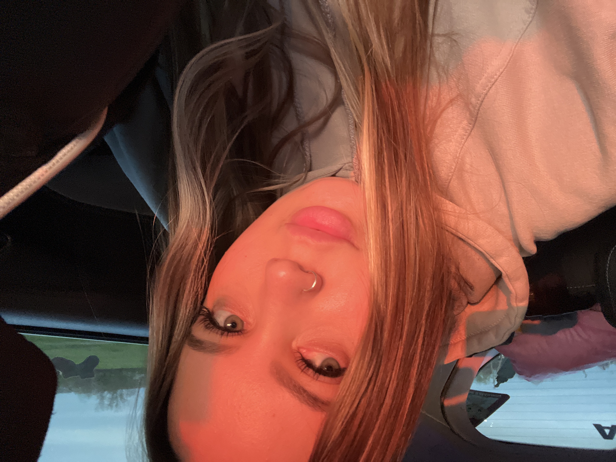 A close-up portrait features a young person with long, blonde and light brown hair, wearing a light-colored hooded sweatshirt, positioned upside down within a vehicle interior. The subject has light-colored eyes, prominent eyelashes, and full, slightly parted lips, with a small silver hoop nose ring in their right nostril.
A strong, warm, reddish-orange light bathes the subject's face and hair from the right, suggesting either sunrise or sunset and creating distinct highlights and shadows. The background reveals the dark upholstery of a car and a rear window. Through the window, a bright, diffused sky and green grassy fields are visible, consistent with an outdoor, rural, or suburban setting in Vyselki, Russia. Indistinct text, possibly an "A," is visible on the car's rear window. The image captures a moment, likely a selfie, emphasizing the subject's features under atmospheric, warm lighting.