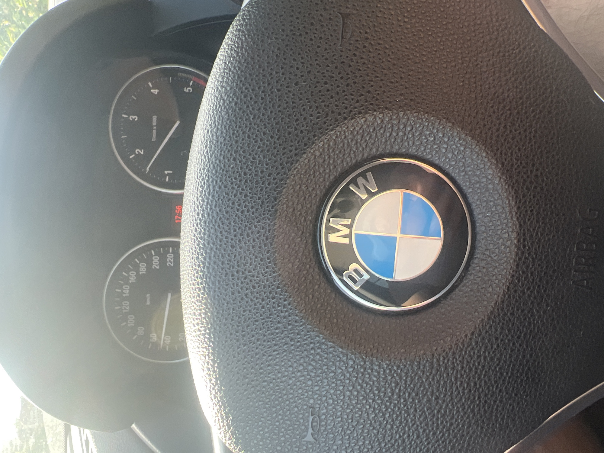 The image depicts the interior of a BMW vehicle, specifically focusing on the steering wheel and the instrument cluster, located in the city of Tsarkva, Bulgaria. The steering wheel, constructed from black textured material, prominently features the BMW logo in its center, which includes the letters "BMW" encircling a blue and white quartered motif. The word "AIRBAG" is visible at the bottom of the steering wheel's central hub, and a horn symbol is present on the left side.
The instrument cluster, partially obscured by bright sunlight, displays two primary dials. The left dial is a speedometer, calibrated in kilometers per hour (km/h), with its needle indicating approximately 50 km/h. The right dial is a tachometer, measuring revolutions per minute (1/min x 1000), with its needle positioned just below 1.5. A digital display, illuminated in red, shows the time as 17:56. Strong sunlight enters the vehicle, creating glare across the instrument panel and the upper portion of the steering wheel. Green foliage is faintly visible through the windshield in the background. No individuals are depicted in the image.