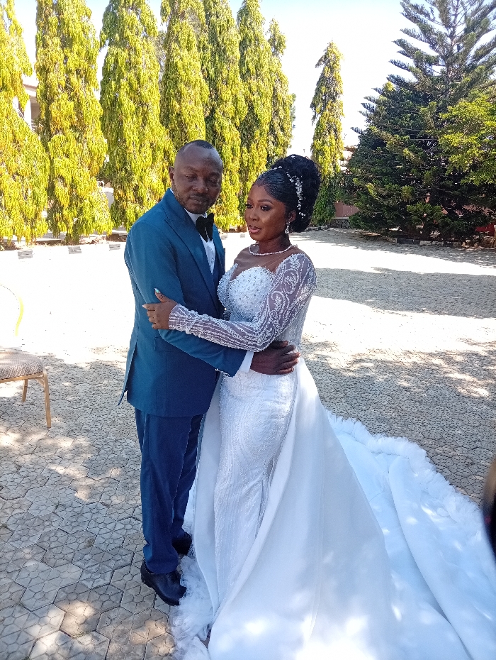 An adult male and an adult female, identified as a couple, are depicted in an embrace outdoors in Lankan, Nigeria. The male, positioned on the left, wears a dark teal suit, white shirt, black bow tie, and black dress shoes, with a patterned garment draped over his left shoulder. The female, on the right, wears a white wedding gown featuring a sweetheart neckline, beaded sheer long sleeves, an embellished fitted bodice and skirt, and a voluminous white train. Her hair is styled in an elaborate updo with decorative elements. Both individuals are looking towards each other with neutral expressions. They are standing on a light-colored paved surface composed of hexagonal or octagonal interlocking pavers. The background features a row of tall, slender evergreen trees with light green foliage under a clear blue sky, indicating bright daylight. A light-colored chair is partially visible on the far left, and additional greenery appears on the right.
