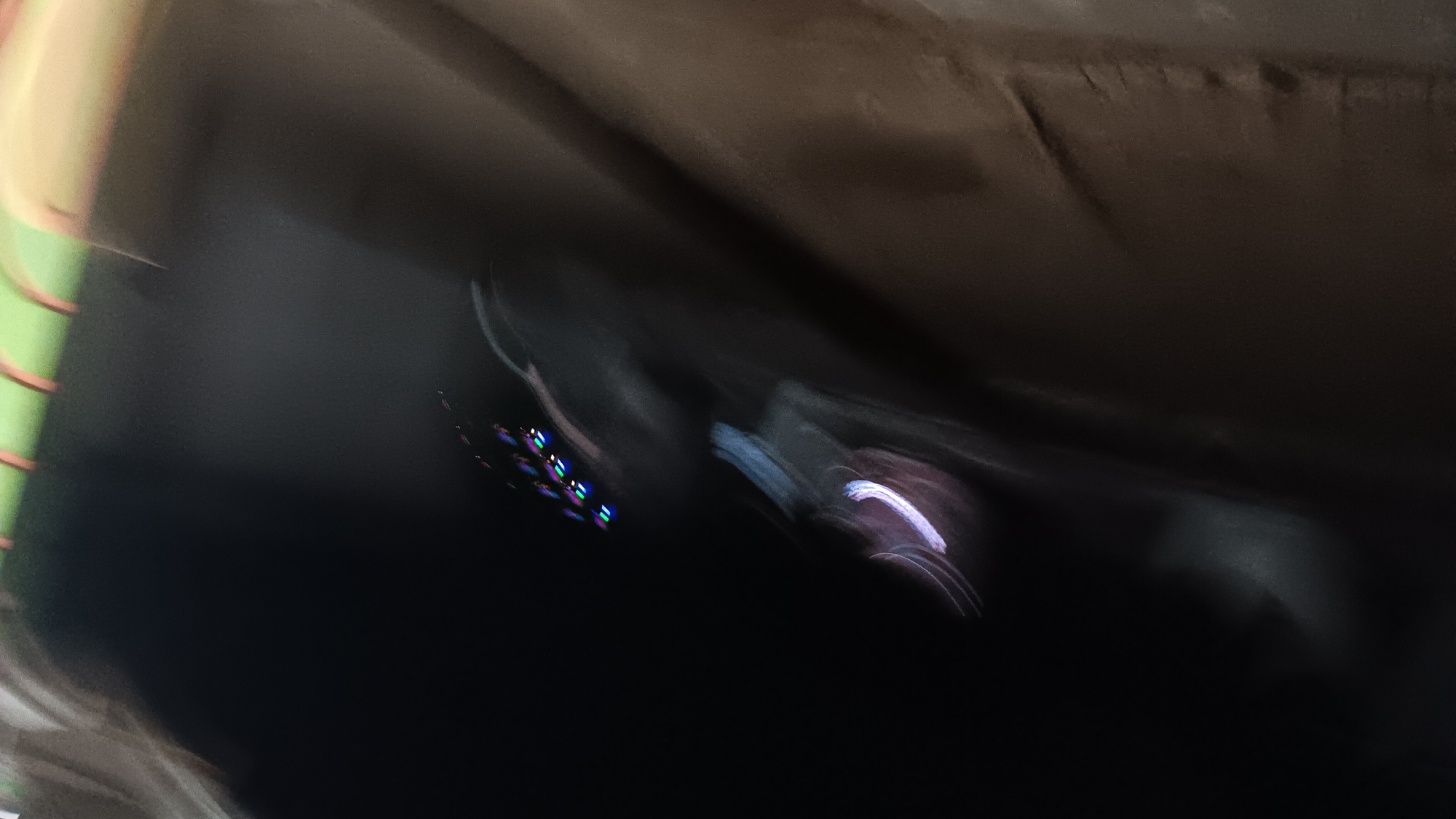 The image is an abstract, blurry, and dark photograph taken in Sonobekel, Indonesia. It appears to be an interior shot with a focus on light trails. In the upper portion of the image, streaks of white and blue light are visible, suggesting movement or a light source. To the right, there is a cluster of small, colorful lights that are also blurred, possibly from a digital display or LED lights. The lower portion of the image is dominated by dark, textured surfaces that are difficult to discern due to the blur and low light conditions. There are no clearly identifiable people or specific objects, and the overall impression is one of motion and atmospheric conditions within an unidentifiable space. The presence of the colorful lights and the dark, enclosed feel might suggest a nighttime setting or an area with artificial lighting, such as a dimly lit entertainment venue or a vehicle interior. The specific location within Sonobekel, Indonesia, is not discernible from the visual information provided.