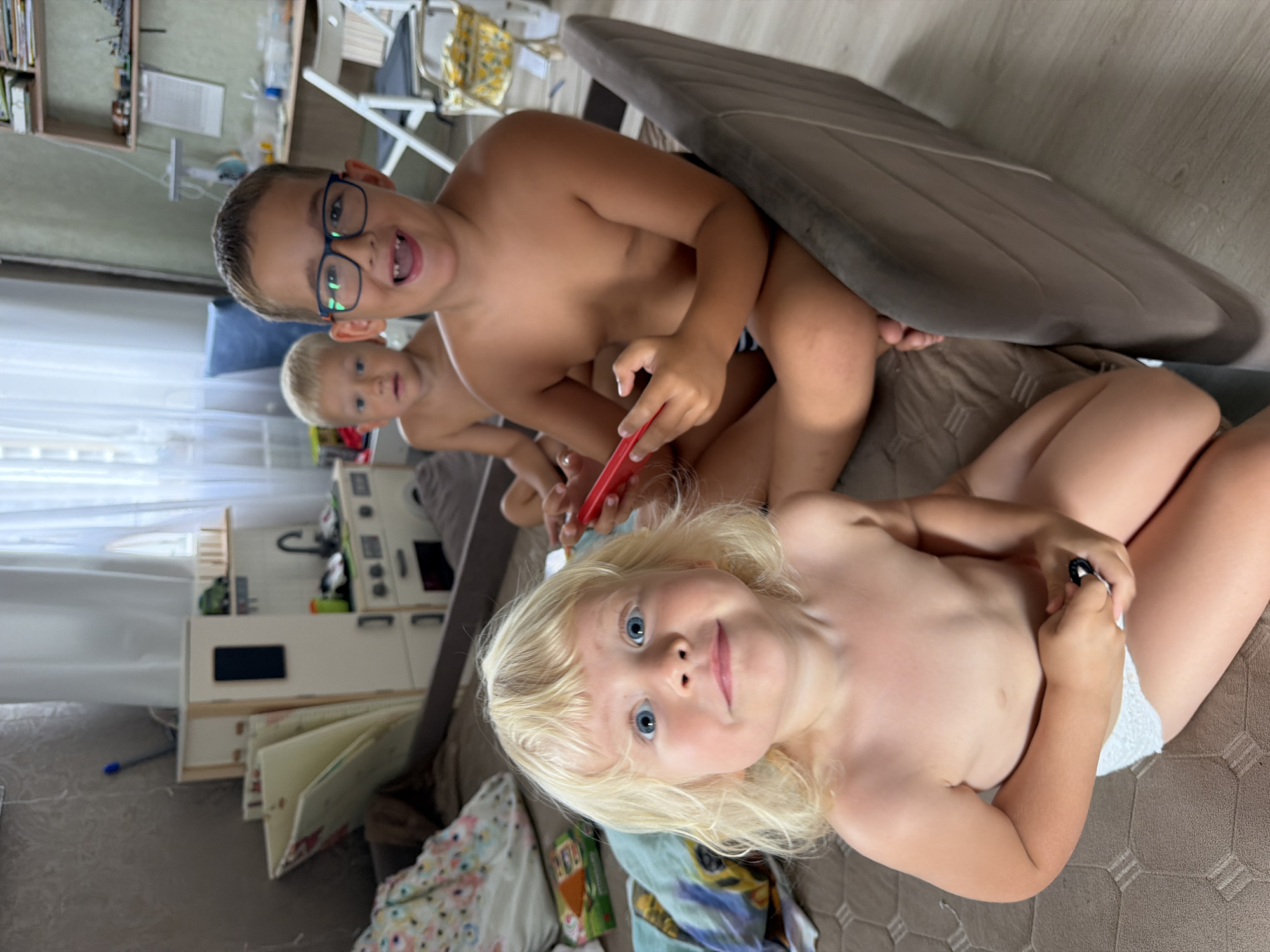The image shows three young children indoors, likely in a home setting in Sochi, Russia. In the foreground, a blonde-haired child with striking blue eyes looks directly at the camera, smiling. This child is wearing a white diaper. Behind them, two other children are visible. The child in the middle has blond hair and is looking slightly away. The child in the foreground on the left is wearing glasses and is looking towards the viewer with an open mouth, appearing excited. This child is holding a red object. The children are seated on a brown, textured surface, possibly a sofa or floor covering. In the background, there are hints of a living space with shelves containing various items, a white kitchen playset, and a window with sheer curtains. The lighting suggests it could be daytime. The overall mood is playful and candid, capturing a moment of sibling interaction and childhood fun.