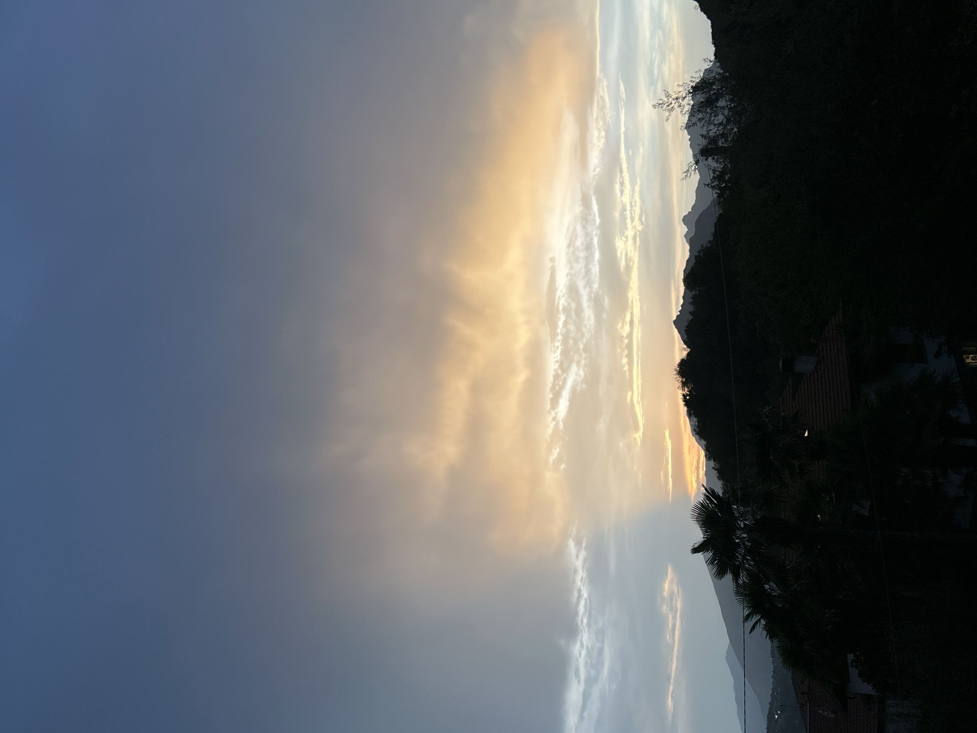 This is a photograph of a sunset over a mountainous landscape. The sky is filled with dramatic clouds, tinged with orange and yellow from the setting sun, contrasting with the deep blue of the evening sky. In the foreground, dark silhouettes of trees and rooftops suggest a residential area nestled amongst the hills. The mountains in the distance are also silhouetted against the vibrant sky. The time of day is clearly sunset, indicated by the warm hues in the clouds and the darkening landscape. The weather appears to be clear, allowing for the full spectacle of the sunset. The location, Calolziocorte, Italy, is hinted at by the presence of tiled rooftops and possibly Mediterranean vegetation like palm trees, partially visible in silhouette. The overall mood is serene and picturesque, capturing the beauty of a natural event. There is no visible text in the image.