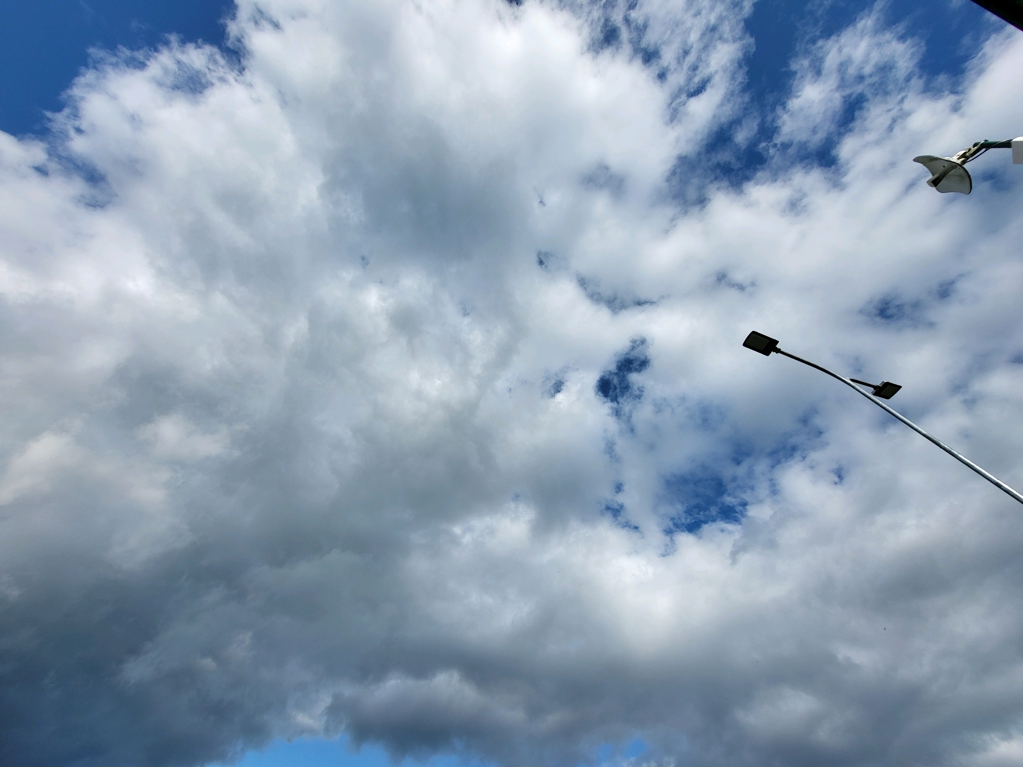 An eye-level, wide shot captures a cloudy sky above Ozolnieki, Latvia. The clouds are varied, with some appearing dense and gray, while others are wispy and white, interspersed with patches of bright blue sky. Two streetlights are visible; one is a modern design with a rectangular head, and the other is a more ornate fixture mounted on a curved arm. The scene is devoid of people or any discernible activity. The environment appears to be an outdoor urban or suburban setting, suggested by the presence of streetlights.