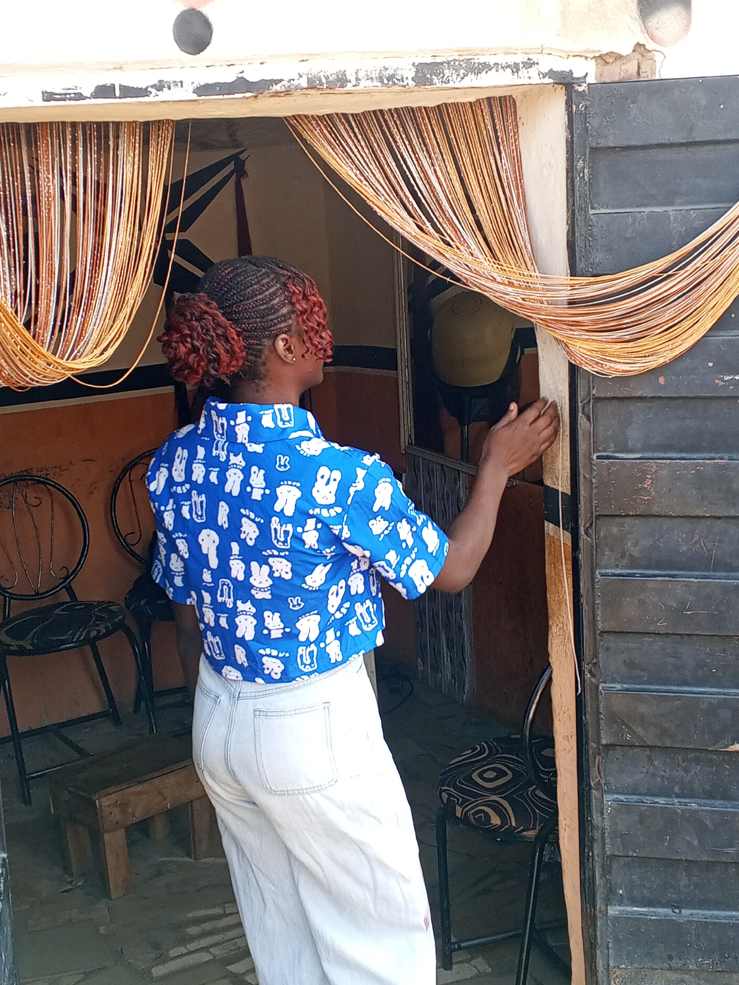 A woman with braided reddish-brown hair is standing in the doorway of a building, possibly a shop, in what appears to be Gwong, Nigeria. She is wearing a blue, short-sleeved shirt with white cartoon-like figures and white pants. Her back is to the viewer, and she is reaching out with her right hand toward a dark-colored, louvered door. The doorway is adorned with beaded strands, and inside, there are chairs and what looks like a salon-style dryer. The building has a rustic appearance, and there is a small wooden stool near the doorway. The floor is tiled.