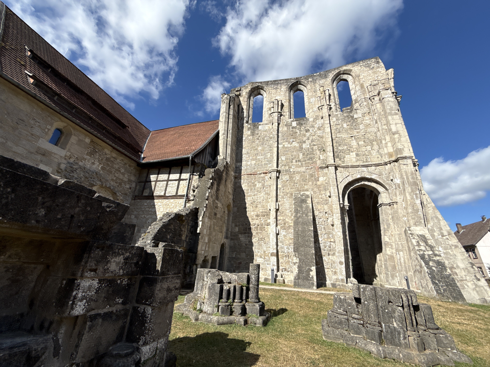 The image shows the exterior of the Walkenried Abbey, a former Cistercian monastery in Walkenried, Germany. The abbey is partially ruined, with stone walls and arched windows visible. In the foreground, there are weathered stone fragments scattered on the grassy ground. The sky is blue with scattered white clouds. To the left, a section of the abbey with a red tiled roof is visible, appearing to be more intact. The overall scene depicts a historical site under a bright, sunny sky.
