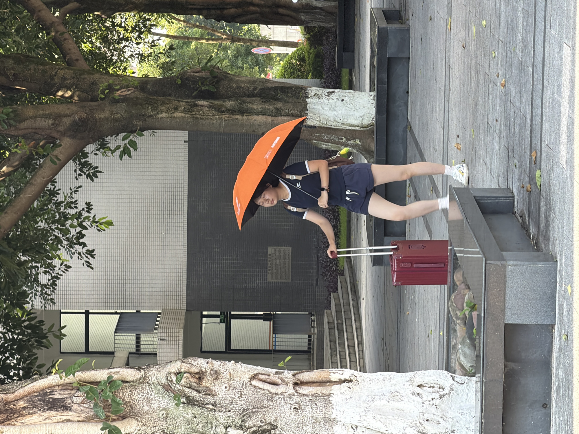 A young woman walks on a paved walkway, holding an open orange umbrella over her head and a red suitcase with a telescopic handle. She is wearing a dark blue t-shirt with white trim, dark blue shorts, white socks, and white sneakers. She has a backpack on her shoulders. She appears to be on the grounds of a building, possibly an educational institution, with trees and a tiled facade visible in the background. The scene is set outdoors during daylight. The location is identified as Jiangjin, China.