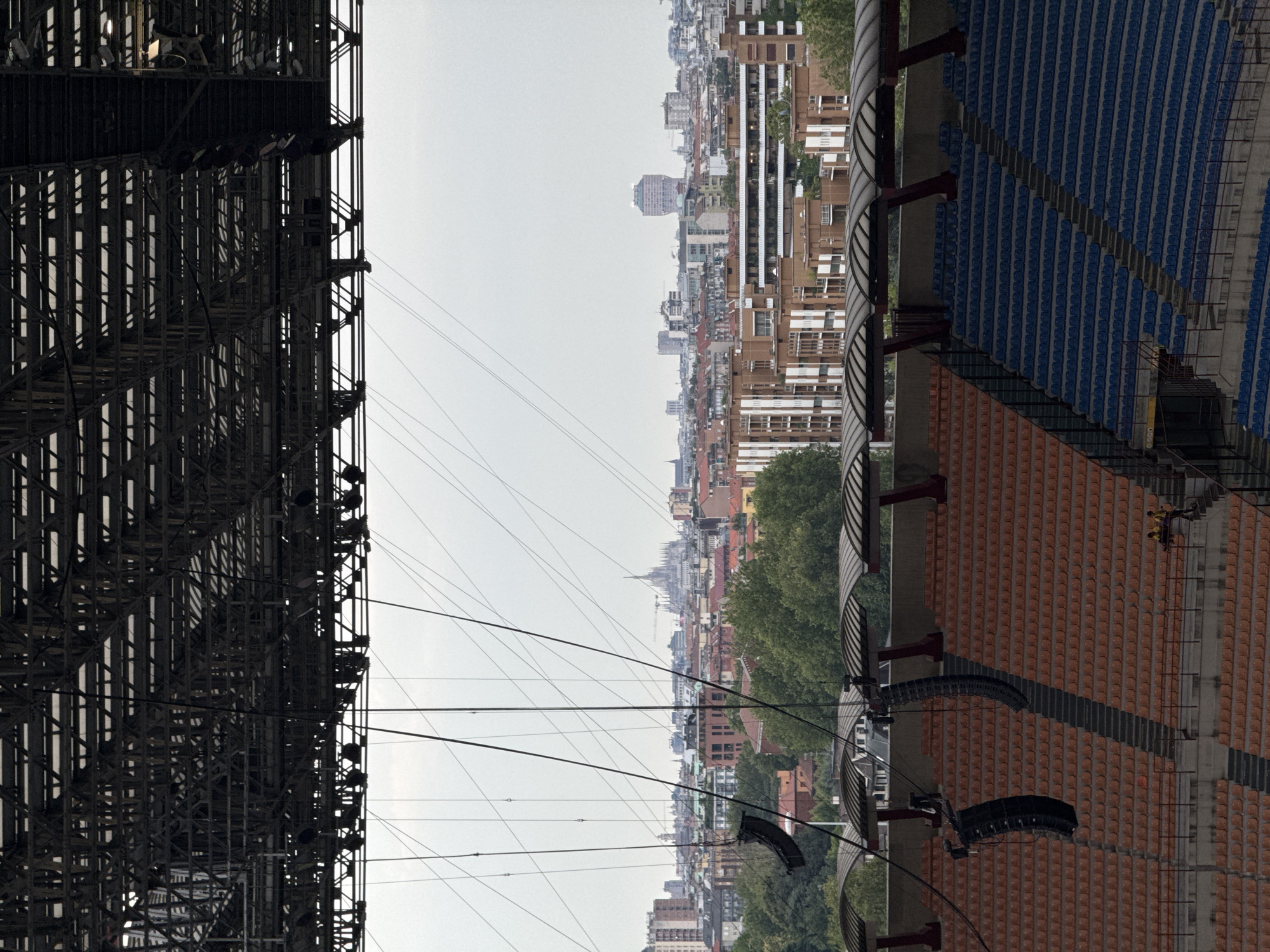 The image shows a view from an empty stadium in Milan, Italy, overlooking the city skyline. In the foreground, rows of orange and blue stadium seats are visible. Above the seats, large speakers are suspended from a complex metal structure that also supports the stadium roof. In the background, the Milan cityscape unfolds, featuring various buildings and a prominent distant cathedral. The sky is overcast. Two individuals are seated on a staircase on the right side of the image.