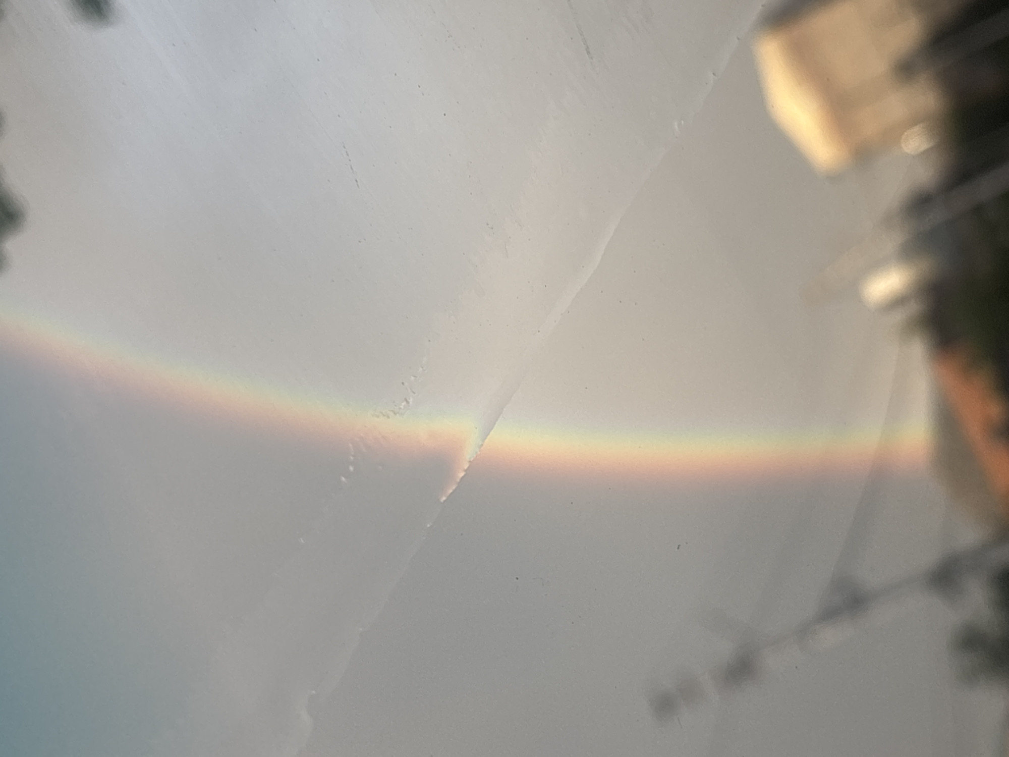 The image shows a rainbow refracted through a dirty windshield, likely a car window, capturing the colors of the rainbow in a horizontal arc. The rainbow is blurred due to the quality of the glass and the camera focus. In the background, out-of-focus, are blurred buildings, trees, and electrical lines, suggesting an urban environment. Based on these details and the prompt, the location is Tbilisi, Georgia, but without more specific details, the exact location within the city cannot be determined. There are no people visible in the image.