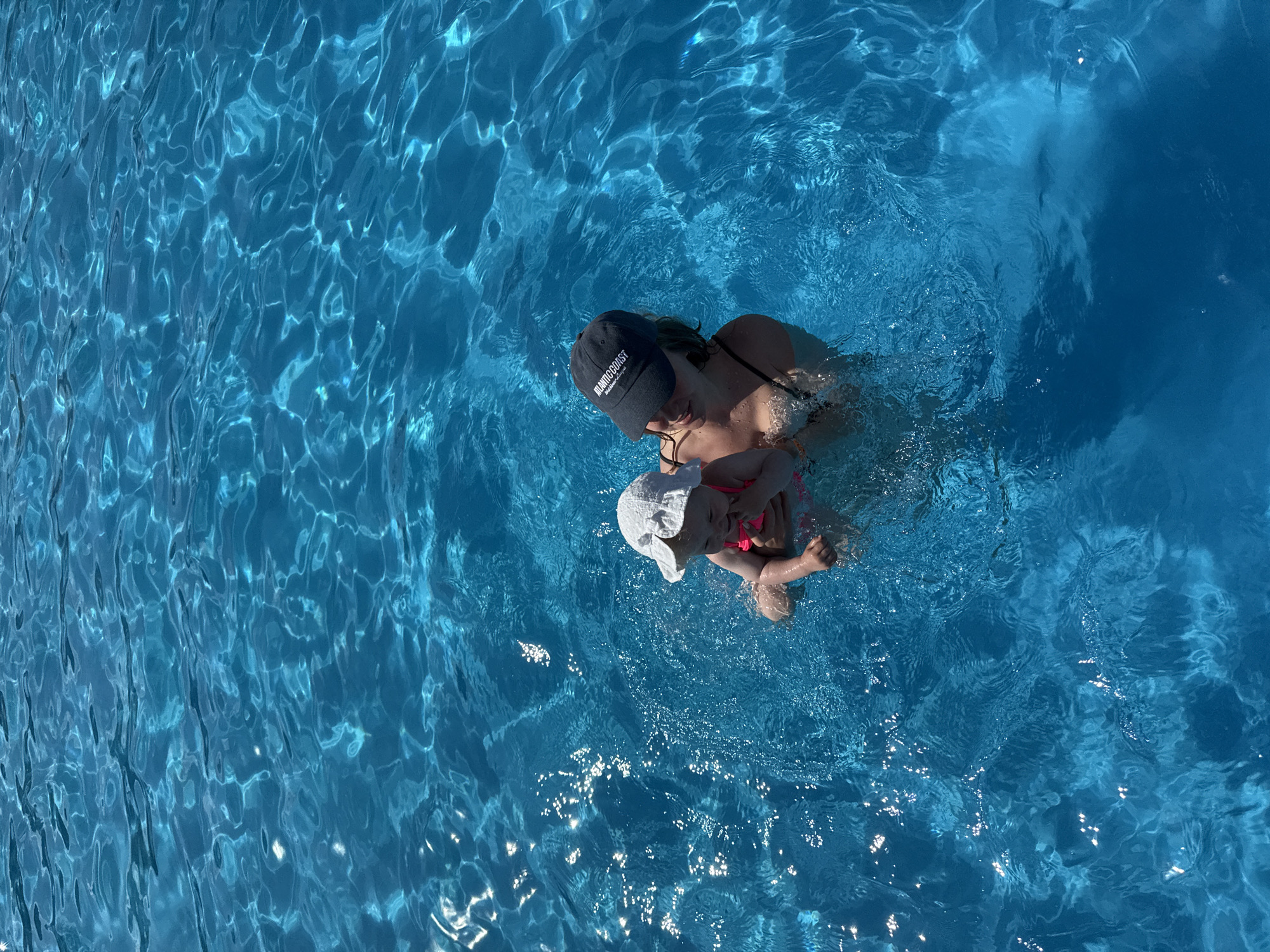 A person and a baby are in a swimming pool. The person is wearing a black baseball cap with white lettering and a black swimsuit. The baby is wearing a white sun hat and a pink swimsuit. The water is a vibrant blue and reflects sunlight. The scene is a bright, sunny day. The location is the city of Koloksha, Russia.