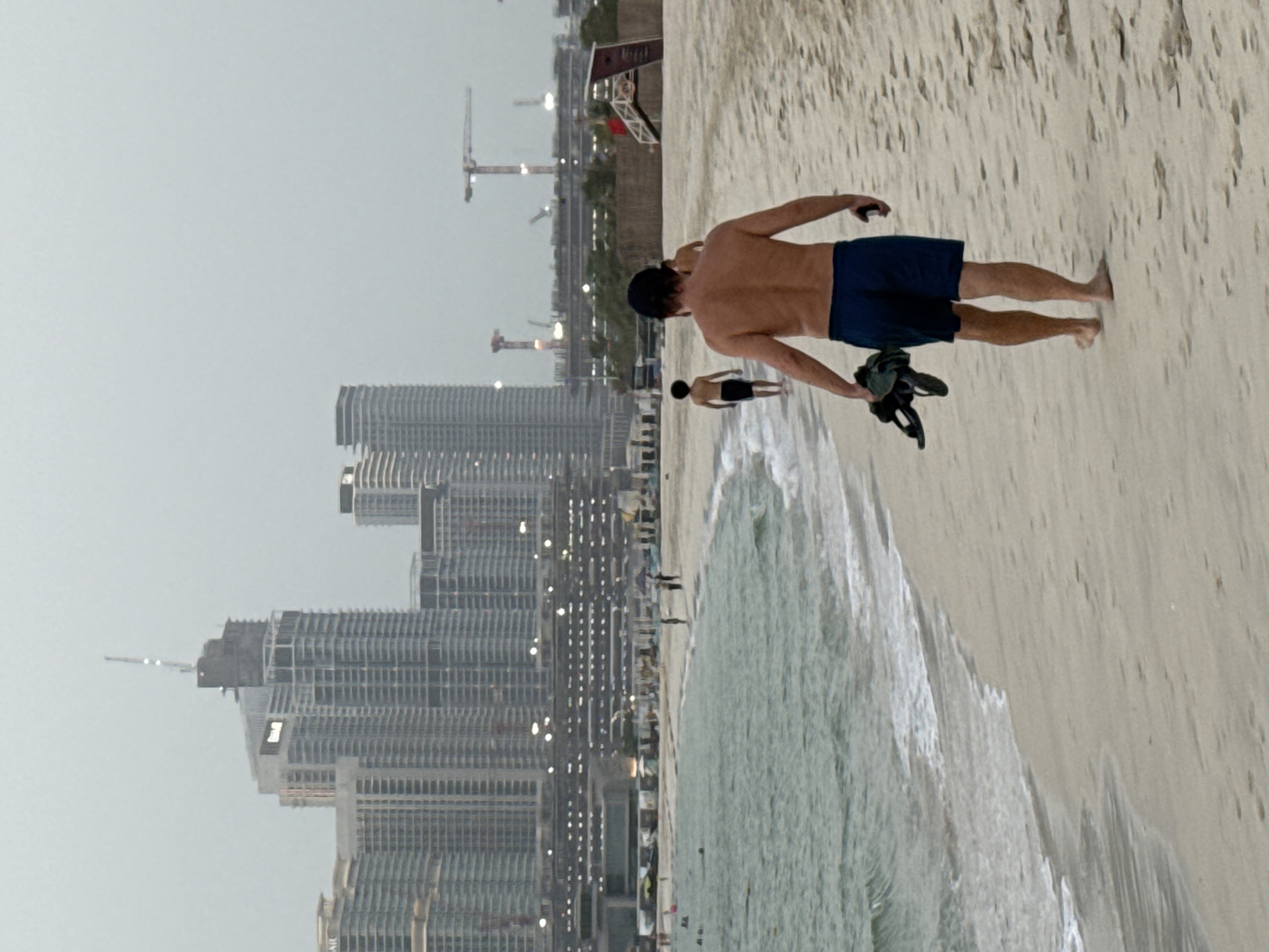 The image captures a beach scene in Dubai, United Arab Emirates, likely Jumeirah Public Beach, given the prominent skyline. In the foreground, a shirtless man wearing dark blue swim trunks walks away from the camera along the sandy shore, carrying a pair of sandals in his left hand. To his left, the ocean laps gently at the beach with small waves. In the mid-ground, a younger person, also shirtless and wearing dark swim trunks, walks further down the beach, also away from the camera. In the background, a dense cluster of high-rise buildings forms the characteristic Dubai skyline, with construction cranes visible amongst them, indicating ongoing development. The sky is overcast, casting a muted light over the scene. Further down the beach, other beachgoers are visible, some near the water's edge and others further inland. A lifeguard tower with a red flag is also present.