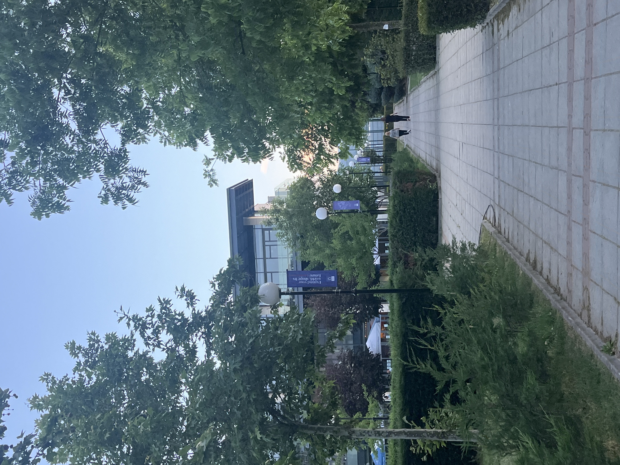 The image shows a walkway in what appears to be the campus of the American University in Bulgaria, located in Blagoevgrad, Bulgaria. The walkway is paved with light grey rectangular stones and is bordered by manicured green hedges and trees. On either side of the walkway are lampposts with spherical white lamps. Banners with the university logo and the words "Expand your world. Shape its future." are attached to some of the lampposts.
In the background, a modern building with a mostly glass facade is visible through the trees. Two people can be seen walking in the distance along the walkway. The sky is light blue with some white clouds visible through the trees. The scene suggests a peaceful and well-maintained campus environment.