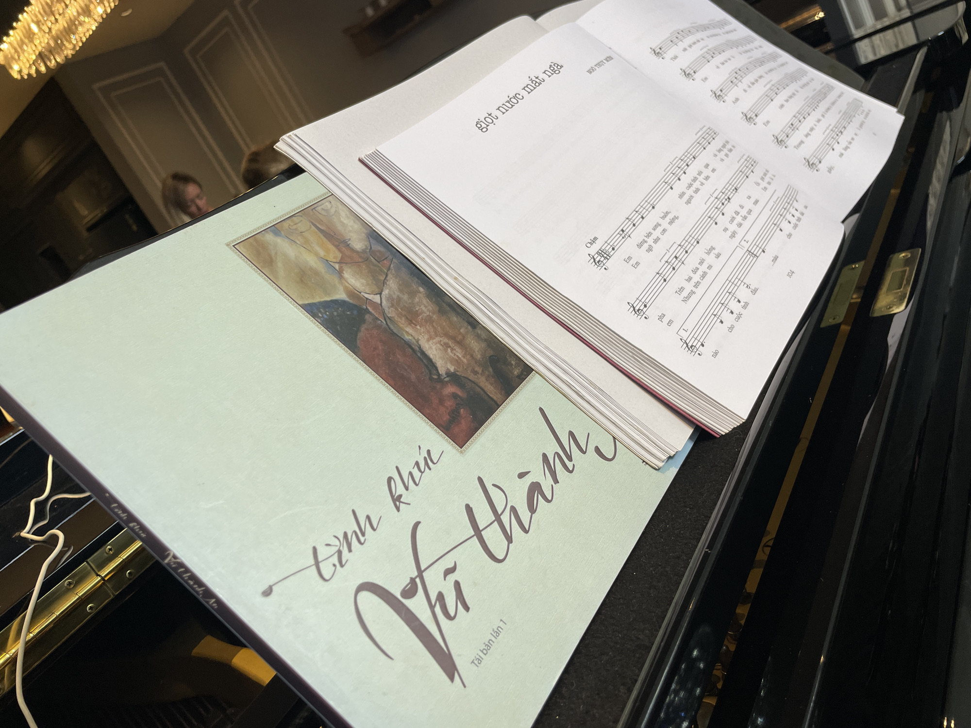 An open sheet music book rests on a black piano, with a light green book titled "Tình Khúc Vũ Thành" placed in front of it. The sheet music book is open to a page with Vietnamese lyrics and musical notation. The green book features an abstract painting on its cover. In the background, a chandelier with multiple lights hangs from the ceiling, and a blurred image of a person is visible in a reflection. The overall scene suggests a musical setting, possibly a performance or practice session in a dimly lit room. The city of Xuân Trường - Đà Lạt, Vietnam is not explicitly depicted or identifiable within the image.