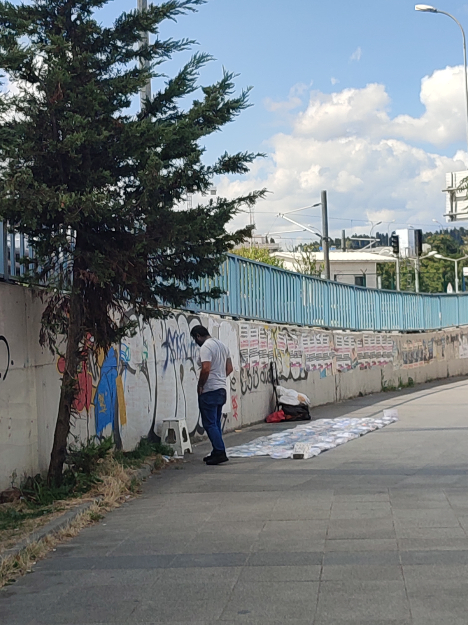 A man wearing a white t-shirt and blue jeans stands beside a concrete wall covered in graffiti and posters, looking down at a collection of CDs laid out on the ground. He is positioned near a small, white stool. To his left, a tree with dark green foliage grows upwards. Behind the wall, a blue railing runs horizontally across the scene, with buildings and trees visible in the background under a sky with scattered clouds. The CDs are arranged in rows on the paved sidewalk, suggesting a possible street vendor or display. The overall setting appears to be an outdoor urban environment.