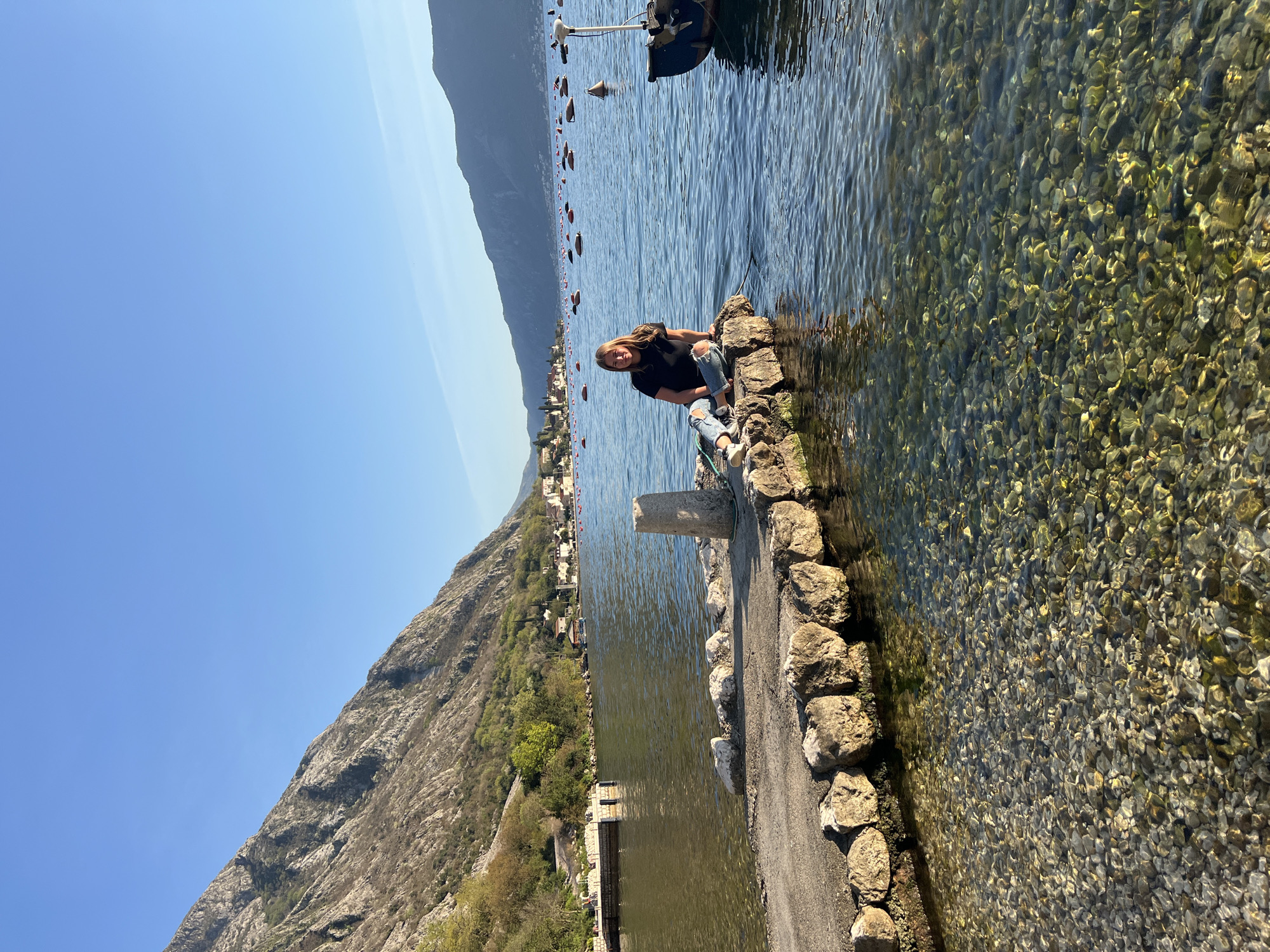 A medium-shot captures a woman seated on a rocky embankment at the edge of the water in Donji Orahovac, Montenegro. She wears a dark t-shirt and ripped jeans with white sneakers. The water is clear, revealing small stones beneath the surface. In the background, a mountain slopes towards the city and the sky is blue. Buoys and a boat are visible further out on the water.