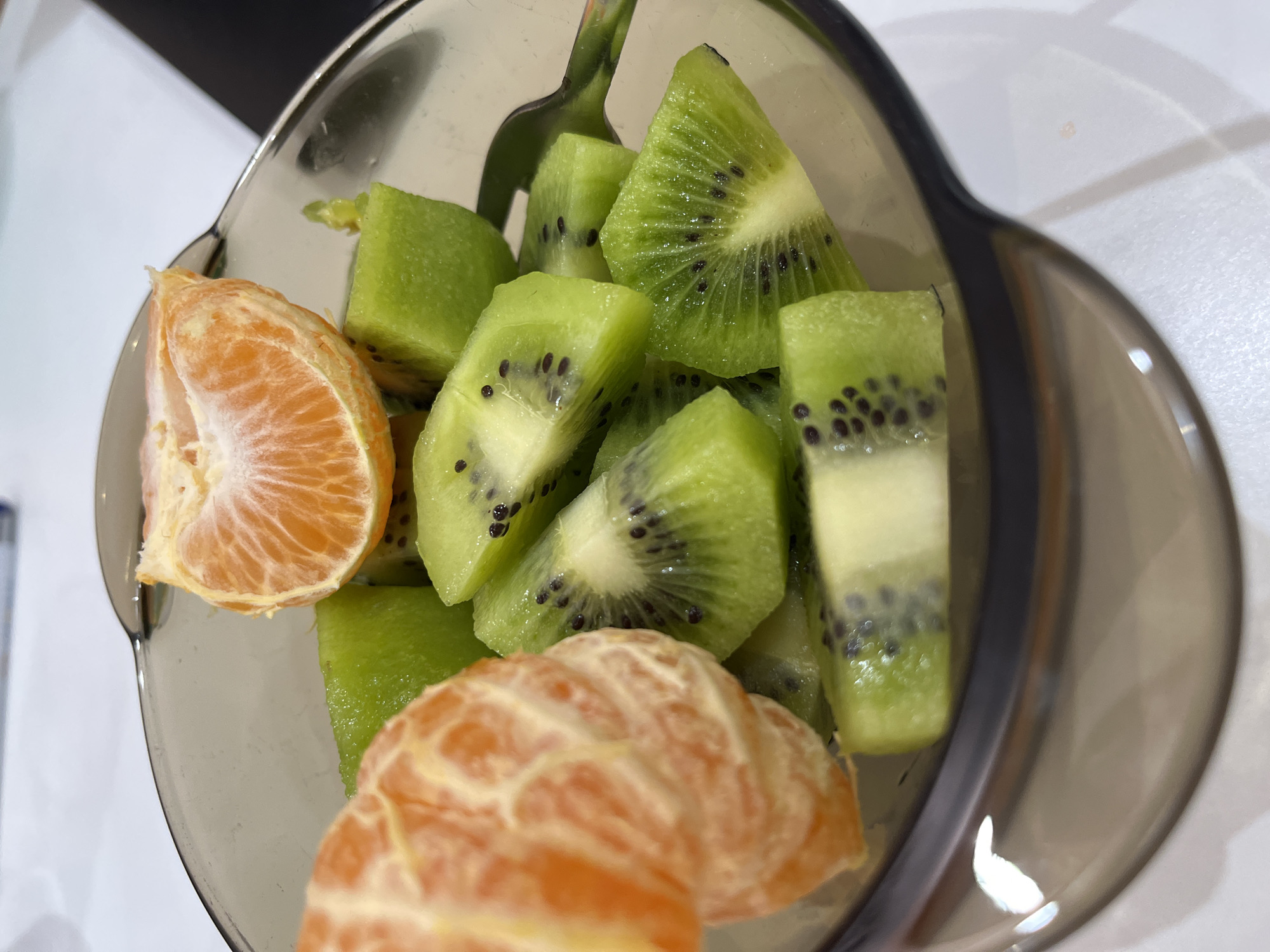 This image presents a close-up, high-angle view of a fruit bowl, located in Pernik, Bulgaria. No individuals are depicted.
The central subject is a dark-tinted, translucent bowl, likely made of glass or plastic, filled with prepared fruit. The contents include numerous pieces of bright green kiwi, peeled and cut into irregular wedges, clearly displaying their white central cores and small black seeds. Several peeled segments of mandarin orange, vibrant orange in color, are interspersed among the kiwi pieces, with one larger peeled section visible on the left and a single segment prominently positioned at the top-center. A reflective metal utensil, consistent with a spoon or fork handle, is partially visible within the fruit on the right side of the bowl.
The bowl rests on a flat, light-colored surface, exhibiting minor reflections. The background is uniformly light and out of focus, indicating an indoor setting. No specific actions or interactions are observable.