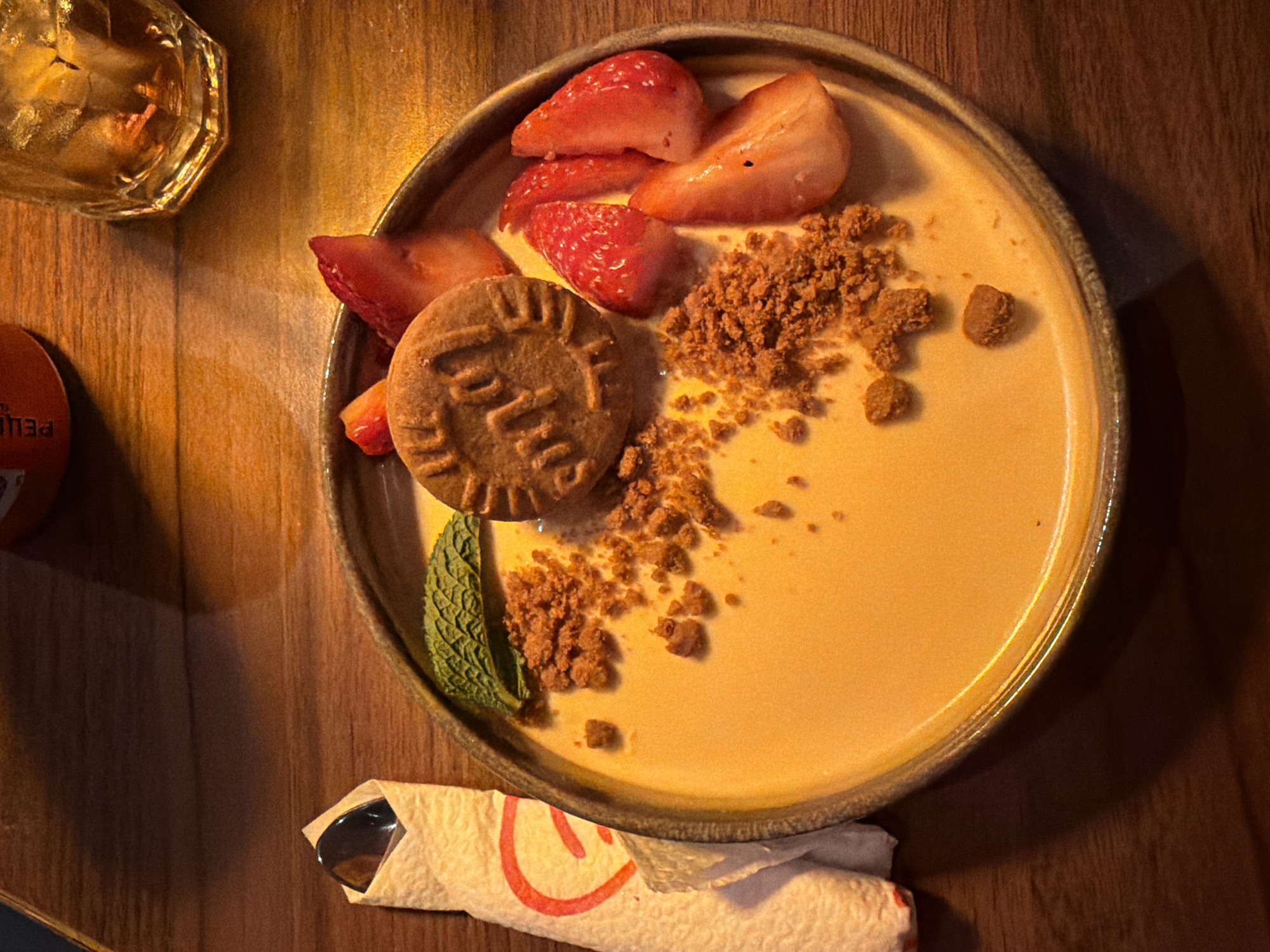 A close-up, overhead shot depicts a dessert plate on a wooden surface, possibly a table. The dessert consists of a circular, tan-colored plate holding a custard-like base. Arranged on top are sliced strawberries, crumbly brown cookie pieces, a single cookie with lettering on it, and a green mint leaf. Next to the plate is a glass of iced tea or a similar beverage and a wrapped utensil. An orange bottle is also visible in the top-left corner of the image. While the image does not provide direct information on the location, the implied setting is likely a restaurant or cafe. Without further data, pinpointing the exact location in Sofia, Bulgaria is impossible.