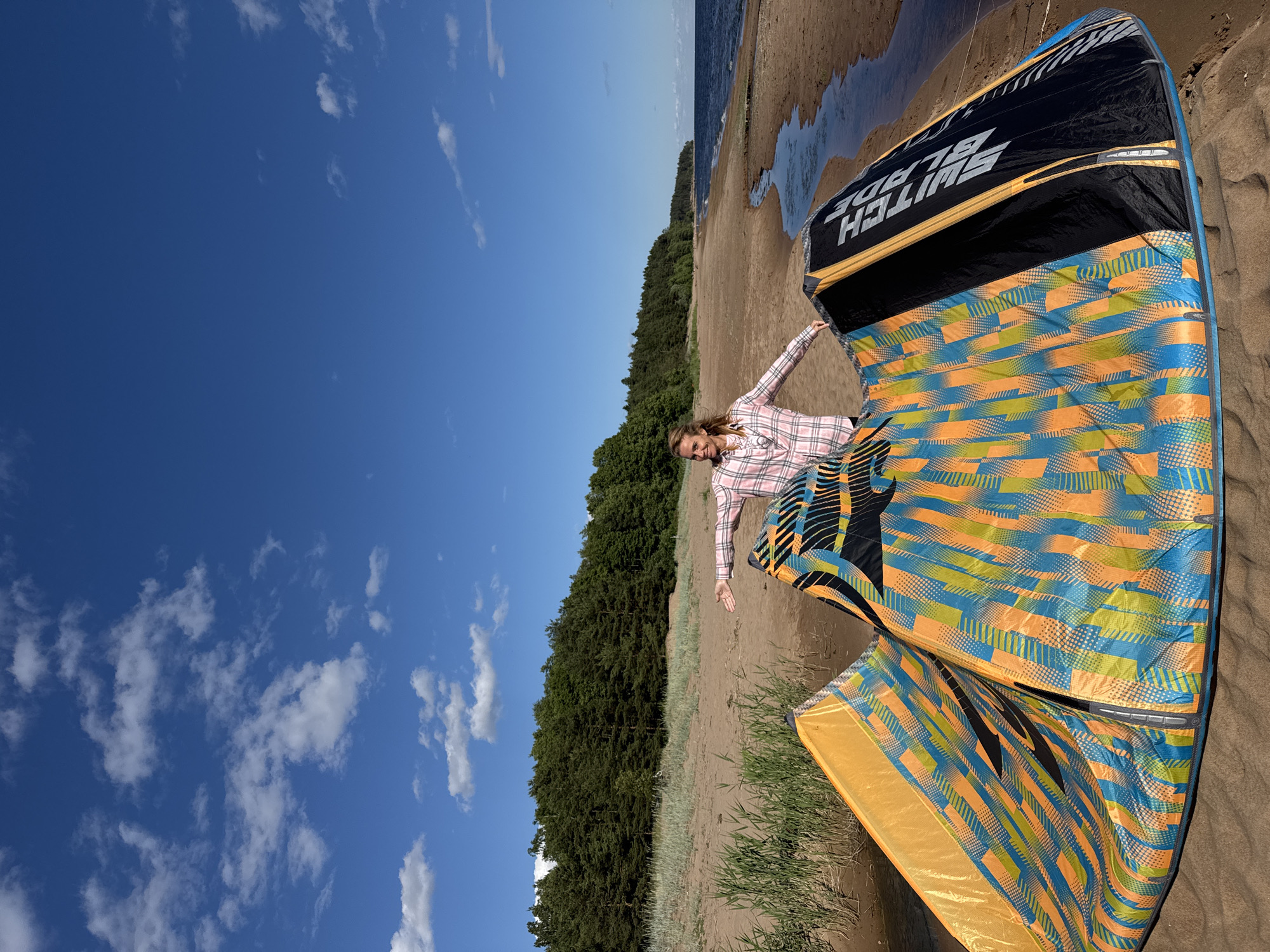 A woman in a pink and white checkered shirt stands on a sandy beach near Zelenaya Roshcha, Russia, with a large, colorful kite spread out on the sand in front of her. The kite features a pattern of orange, blue, and yellow geometric shapes. The woman has her arms outstretched, and she is smiling and looking to her left. Behind her, a line of trees borders the beach, and a calm body of water stretches out to the horizon under a bright blue sky with scattered white clouds. A small stream of water flows into the larger body of water on the right side of the image. The sand is light brown, and there are some green reeds growing in patches along the shoreline. The scene appears to be a sunny day.