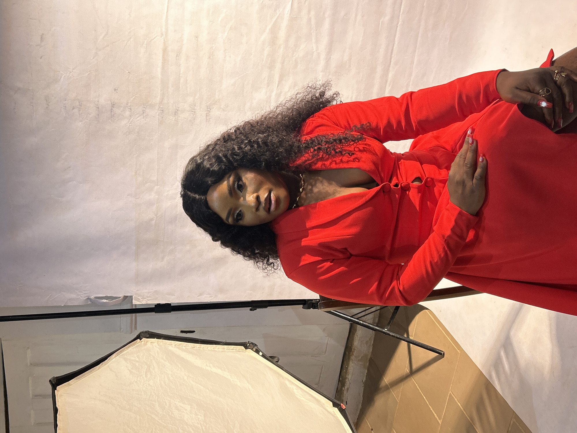 A woman with dark curly hair, wearing a low-cut red dress and gold necklace, is posing in front of a textured white backdrop. Her hands are adorned with rings and patterned nails. A softbox light is positioned in the foreground, illuminating the scene. The setting appears to be a studio or indoor space. There is no indication that this image was taken in Jos, Nigeria.