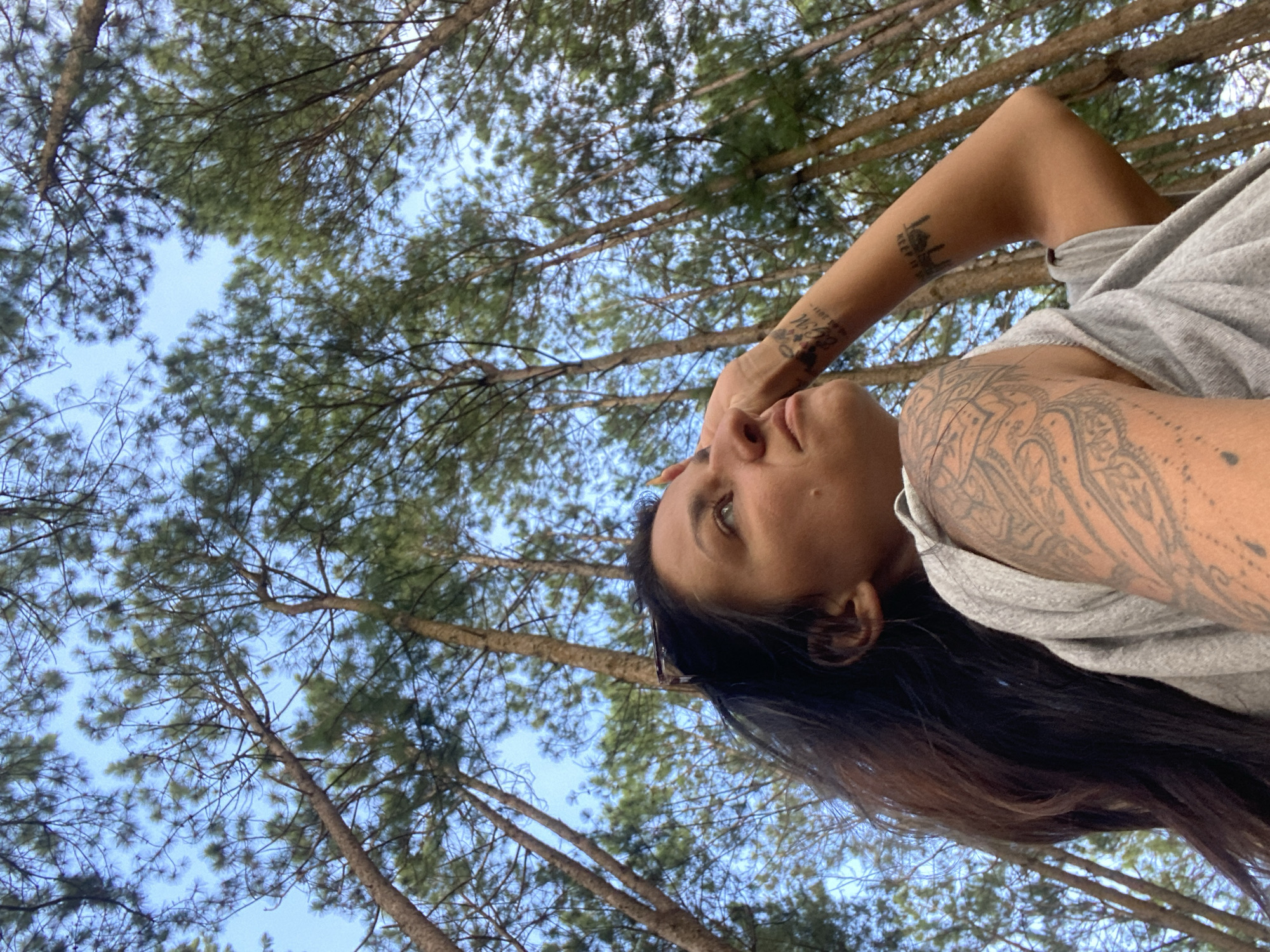 A woman with dark hair and tattoos on her left arm is looking up, with her right hand resting on her forehead. She is wearing a gray t-shirt. The background consists of tall pine trees and a blue sky. The image appears to be taken from a low angle, looking up into the canopy of the trees. The location is specified as Thôn 3, Vietnam.