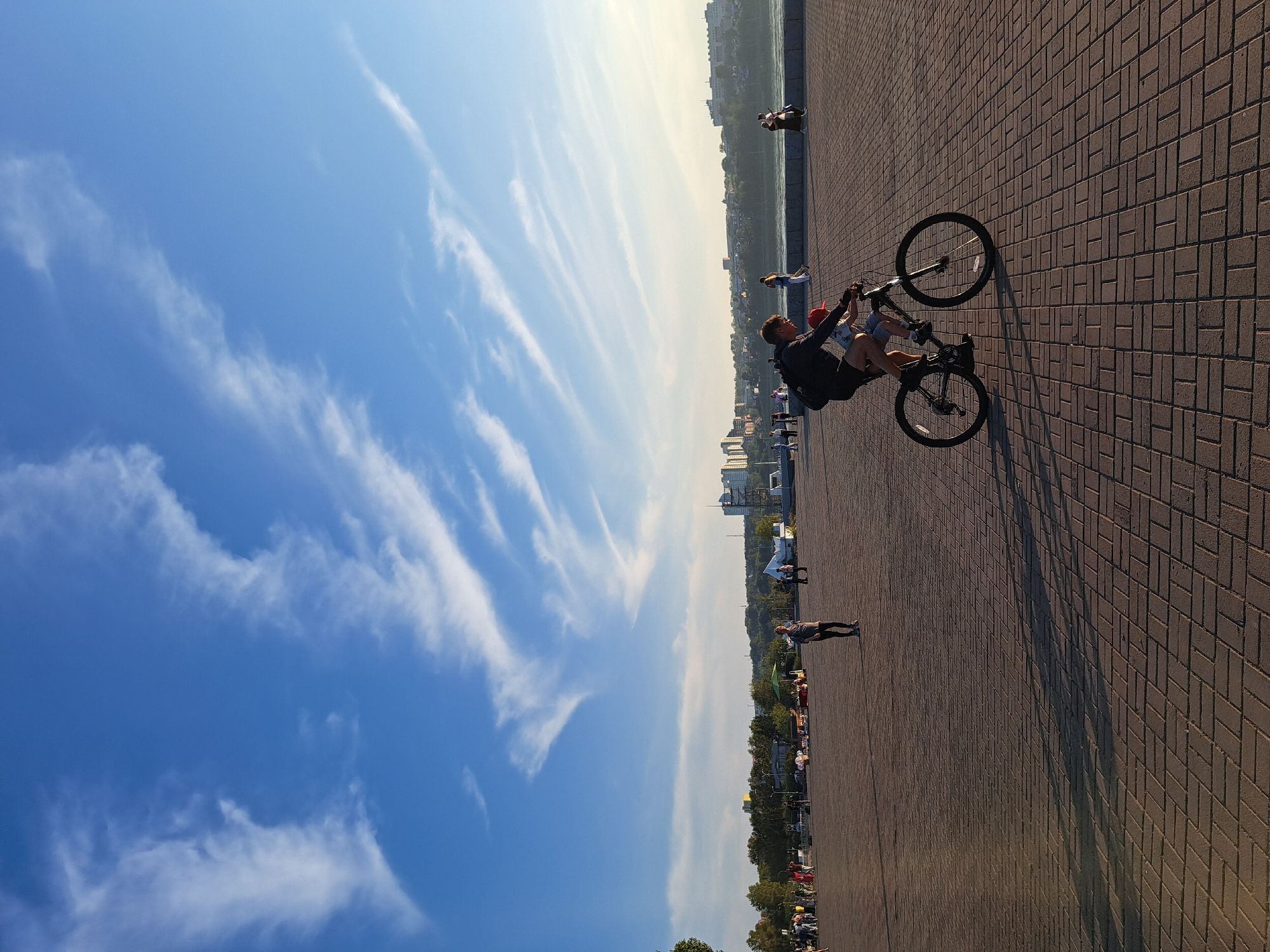 An outdoor scene captures a bright, clear sky with wispy white clouds stretching across the expanse. In the foreground, a man with dark hair wearing a black t-shirt and shorts is riding a black bicycle. A young child, wearing a red cap and blue shirt, is seated on the bicycle's handlebars, facing forward. The bicycle has two black wheels and a silver frame. They are moving across a wide, paved area constructed of reddish-brown brickwork arranged in a herringbone pattern. The paving casts long shadows, indicating the sun is low in the sky, likely during late afternoon or early evening.
In the middle ground, a paved promenade stretches towards a body of water, likely a river or lake, bordered by a low wall. Beyond the water, a cityscape is visible, characterized by buildings of various heights, including some taller structures with modern architecture and numerous trees dotting the landscape. A line of people is scattered across the promenade and on the far bank, some walking, others gathered in small groups. Some stalls or vendor carts are present on the left side of the promenade.
The image appears to be taken from a slightly elevated perspective, looking towards the cityscape. The overall atmosphere is peaceful and suggests a public recreational area. Based on the architectural styles and the general appearance of the landscape and cityscape, this scene is likely located along the Angara River embankment in Irkutsk, Russia, specifically the area near the city center.