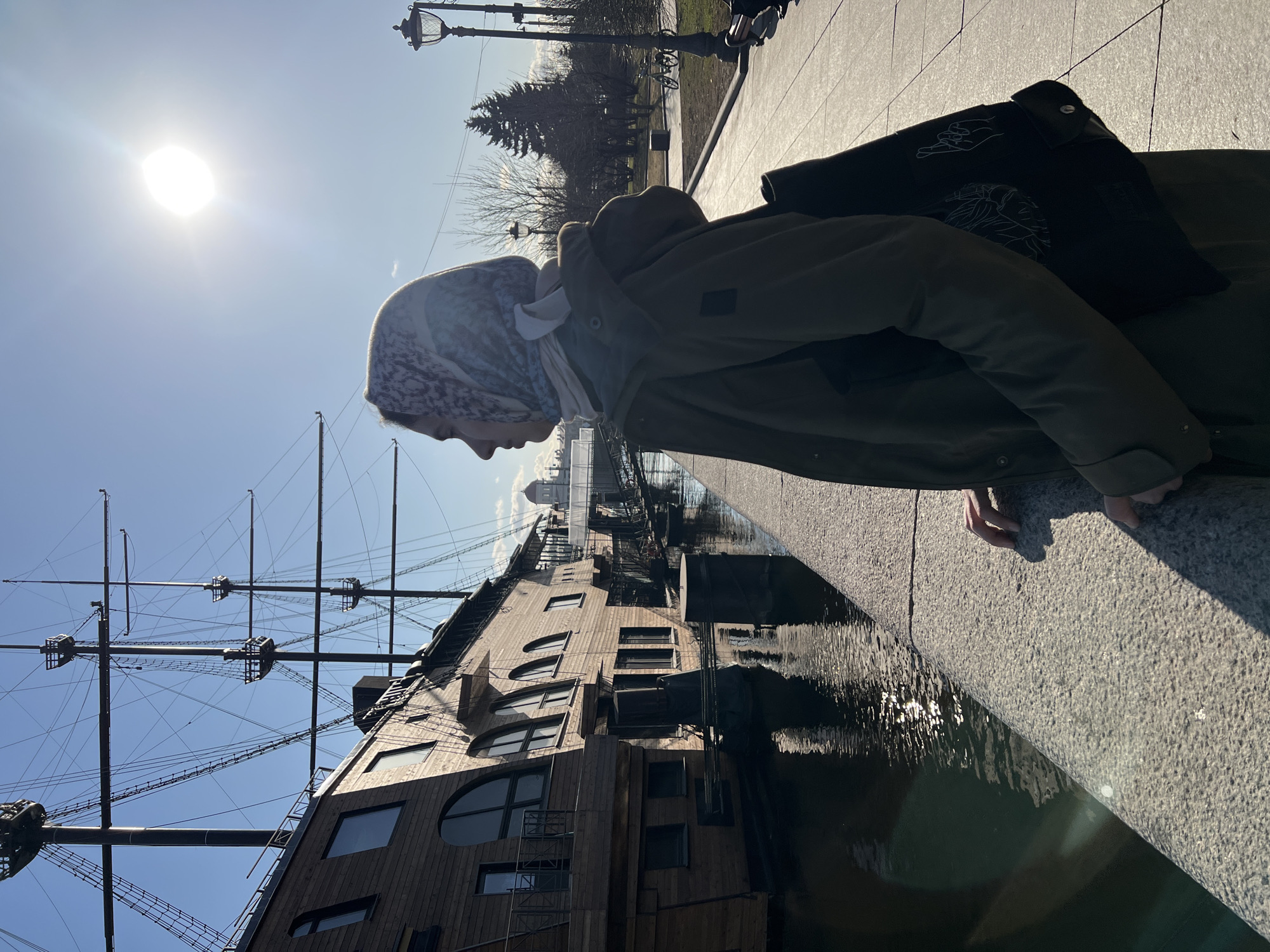 A person is depicted in profile, standing on a grey granite embankment in Saint Petersburg, Russia. The location is the Malaya Neva River, with the "Flying Dutchman" restaurant-ship moored directly opposite the embankment. The subject, a female, is positioned in the right-center foreground, wearing a patterned headscarf, an olive-green coat, and carrying a dark shoulder bag adorned with white line art depicting a face and crossed fingers. Her hands rest on the embankment wall as she looks towards the right.
To the left, the large wooden "Flying Dutchman" ship features multiple masts with intricate rigging extending against a clear blue sky. Its hull is characterized by numerous windows, some arched and some rectangular. The dark water of the river reflects the ship and sky, with a strong glare from the sun.
The background on the right includes a paved pedestrian path, an ornate dark street lamp, and a park area containing bare deciduous trees and dark evergreen conifers. Bicycles are visible parked in the distance. A bridge is partially visible in the middle ground, and a light-colored building with a distinctive dome, consistent with the Peter and Paul Fortress, is discernible on the horizon, behind the ship. The sun is a prominent, bright light source in the upper right quadrant of the sky, casting long shadows.