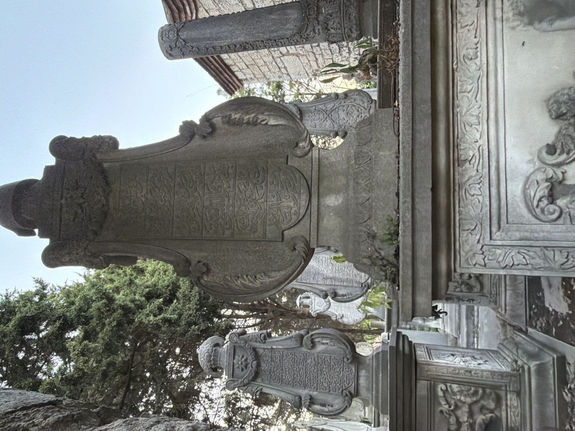 The image is a low-angle, full-shot photograph of gravestones in a cemetery. The primary subject is a large, ornate, grey stone gravestone in the center foreground, with intricate carvings and Arabic script. To the left is another smaller, grey stone gravestone with similar decorative elements and script. In the background, several other gravestones of varying sizes and designs are visible, along with trees and a brick building. The sky is clear and light blue. The overall atmosphere is solemn and quiet. The gravestones suggest this is likely a historical cemetery, potentially in Eyüpsultan, Turkey, an area known for its significant religious and historical sites, including the Eyüp Sultan Mosque and its associated cemetery, which contains many elaborately decorated tombs.
