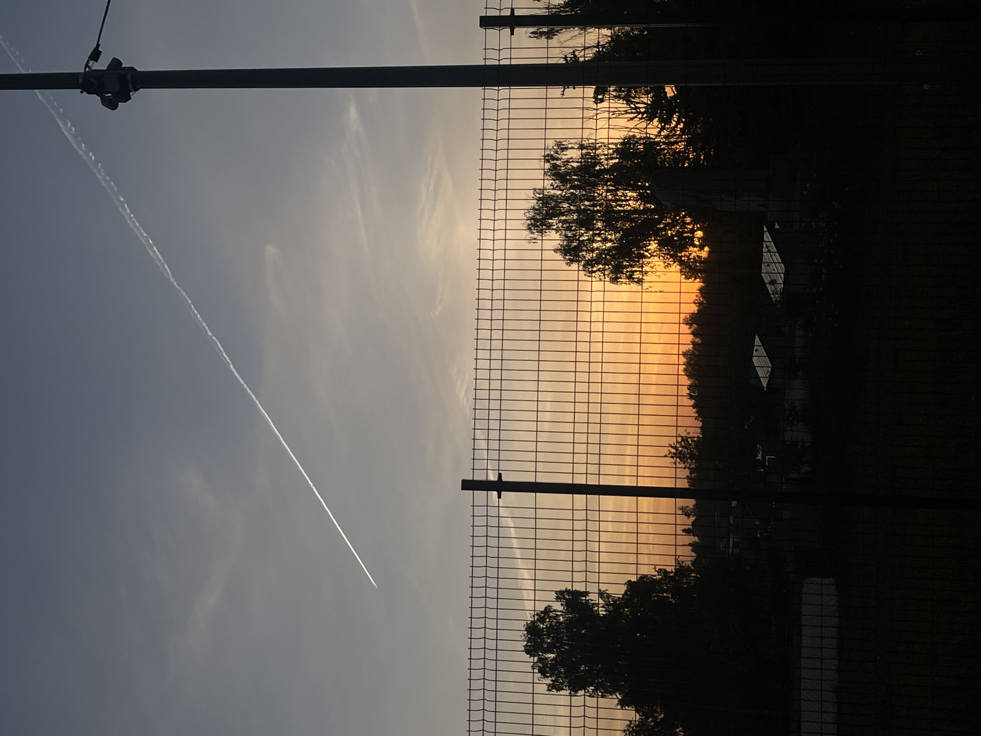 The image depicts a horizontal view of a chain-link fence with supporting posts. To the left of the fence, a vapor trail cuts across the sky. To the right, the setting sun glows behind trees and a structure that appears to be a building. A light post is visible above the vapor trail on the left. The location appears to be Фоки, Russia, based on the metadata. The image does not contain information on people, interactions, or actions.