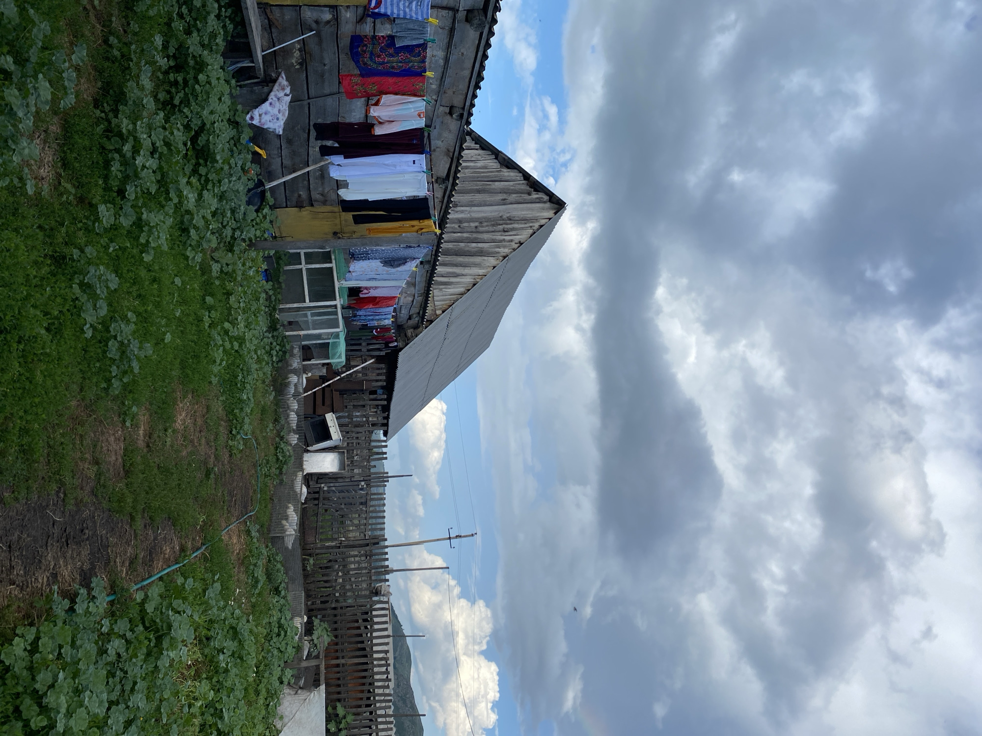 This image depicts a rustic scene in Maloarslangulovo, Russia, featuring a wooden house with laundry hanging on a clothesline in front. The house appears aged, with weathered wooden walls and a corrugated metal roof. Various articles of clothing, including shirts, pants, and a patterned cloth, are spread out, secured by clothespins. A garden path, partially overgrown with greenery, leads towards the house. The surrounding environment includes a wooden fence, a utility pole with wires, and distant hills under a cloudy sky. The overall atmosphere suggests a rural setting, possibly a dacha or a traditional Russian countryside dwelling.